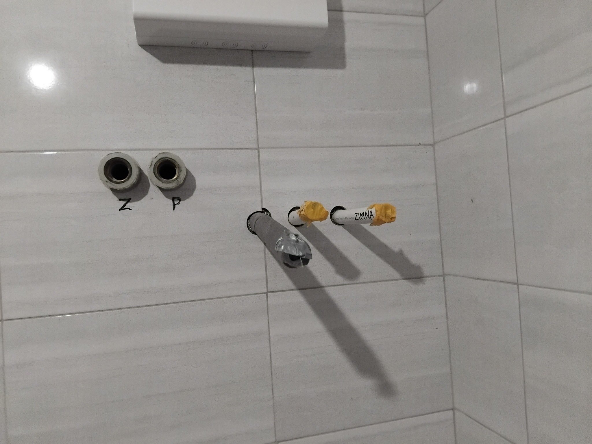 Szare płytki, wystające rury z oznaczeniami 'Z' i 'P' oraz 'ZIMNA', sugerujące instalację wodną w łazience, z widocznym fragmentem białego grzejnika.
