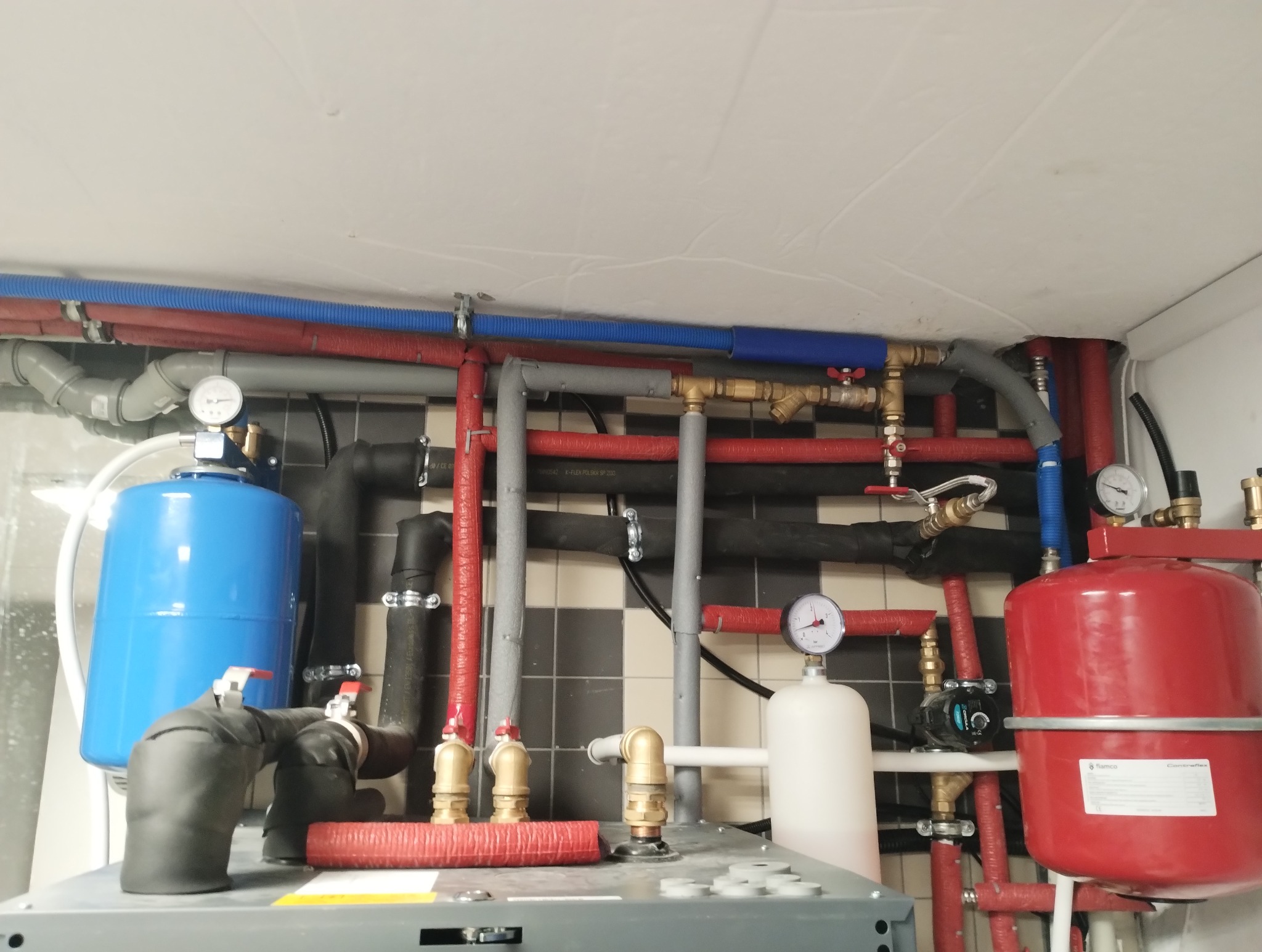 Złożona instalacja grzewcza z rurami w kolorach czerwonym, niebieskim i czarnym, zbiornikami wyrównawczymi: niebieskim i czerwonym, manometrami na tle czarno-białych płytek.