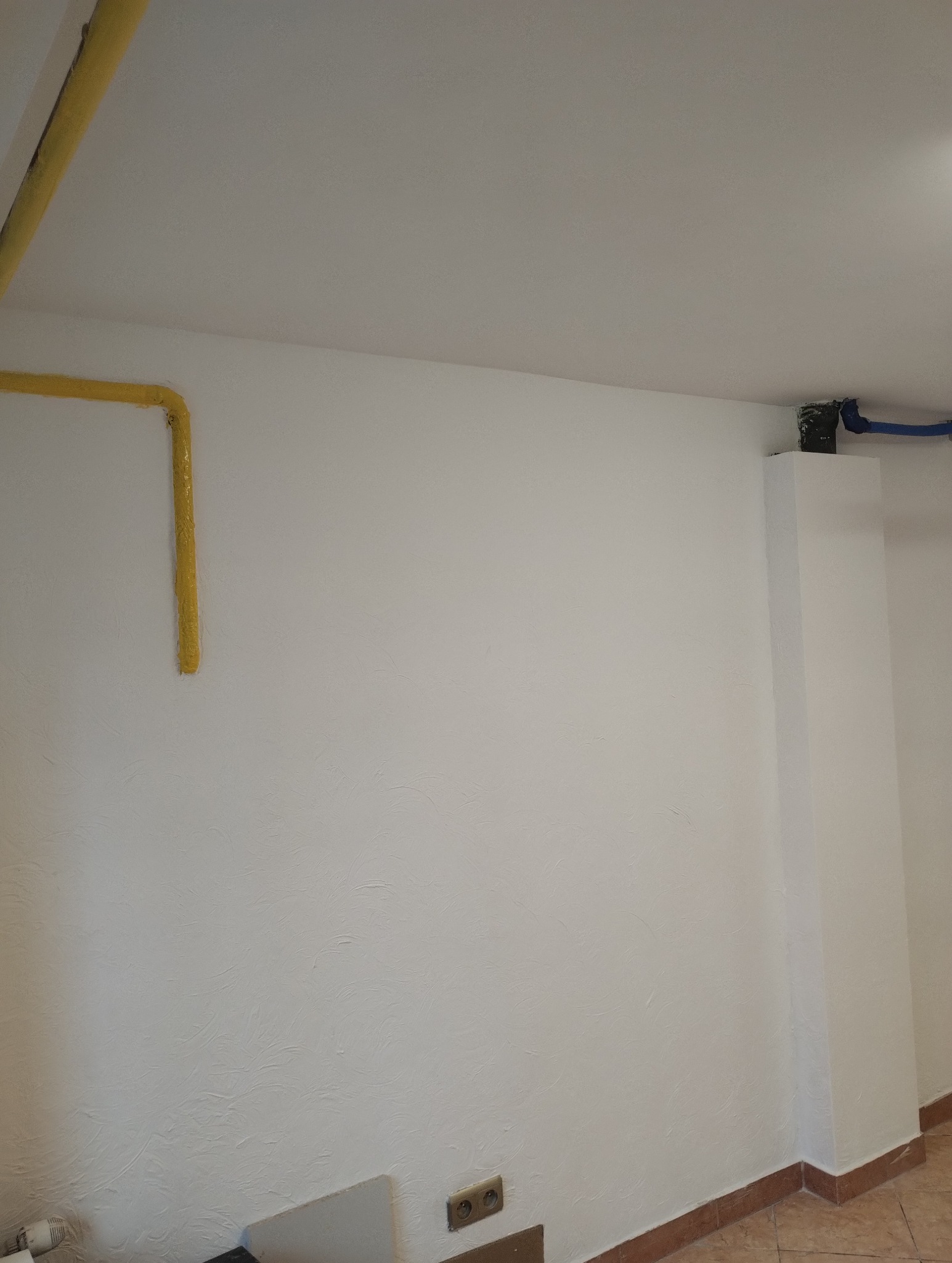 Biała ściana z żółtą instalacją gazową i niebieską instalacją wodną, widoczne gniazdka elektryczne i fragment podłogi. Świeżo po szpachlowaniu.