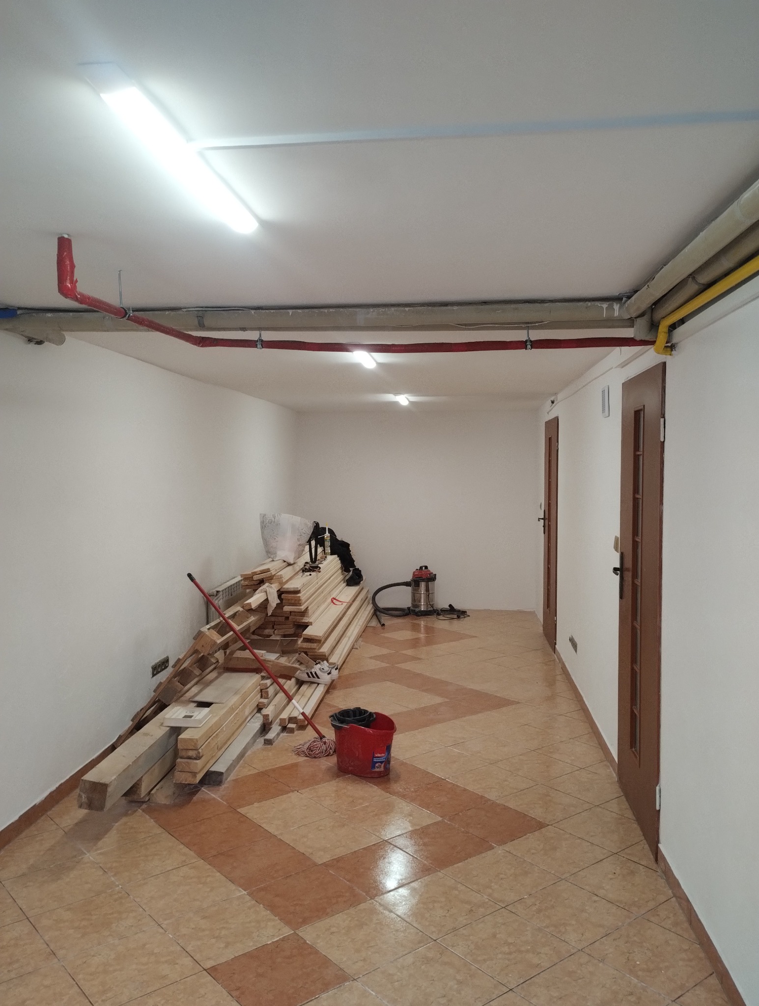 Wnętrze w trakcie remontu: ułożone deski pod ścianą, wiadro z mopem na brązowych płytkach podłogowych. Widoczne rury pod sufitem i dwie drewniane, zamknięte drzwi.