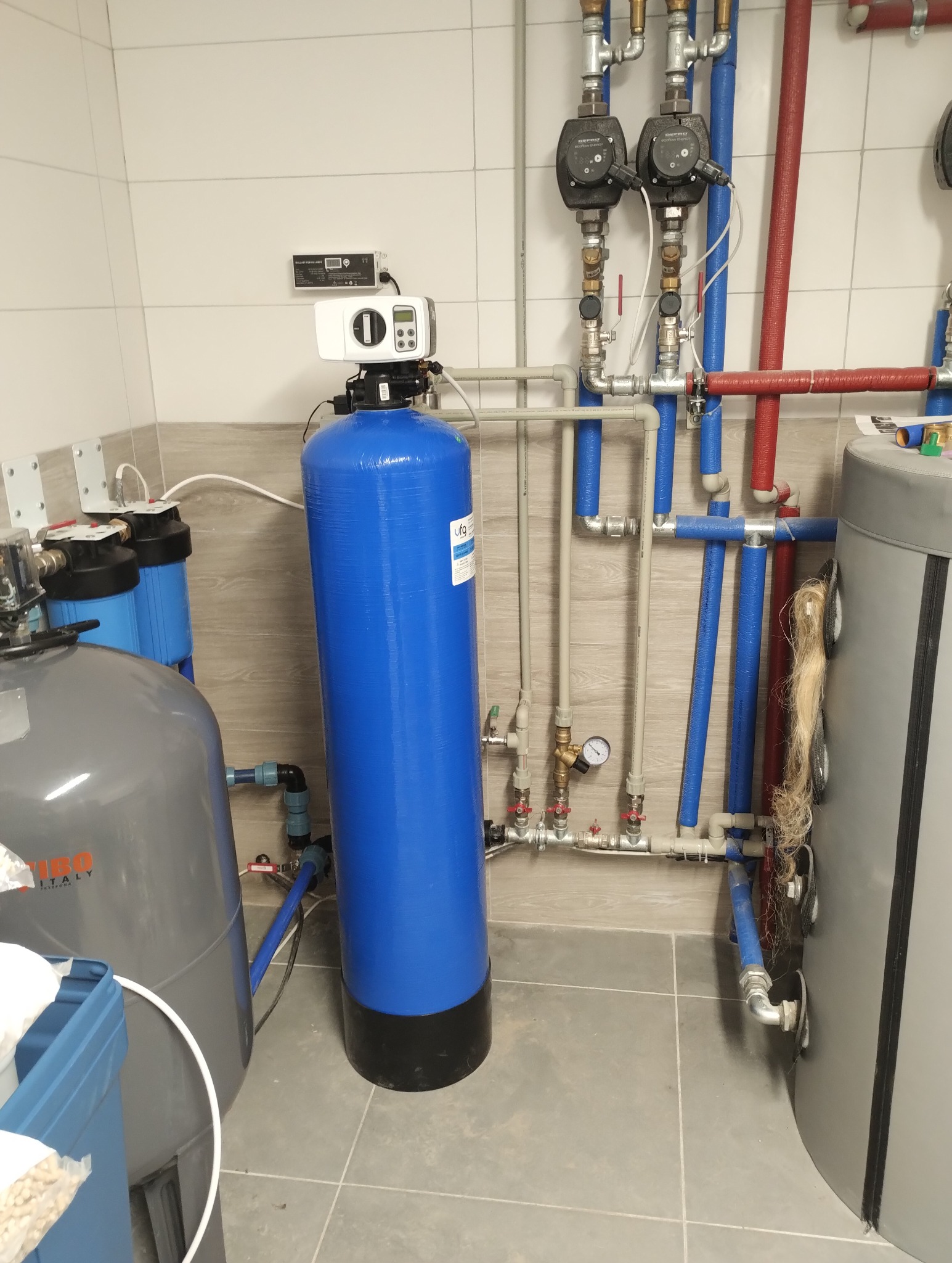 Kompletna instalacja uzdatniania wody w kotłowni: zbiorniki ciśnieniowe, rury w izolacji, zawory i manometry. System zamontowany na płytkach podłogowych.