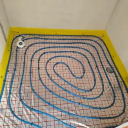 Niebieskie rury ogrzewania podłogowego ułożone spiralnie na izolacji z żółtą taśmą brzegową, widok z góry w surowym wnętrzu.