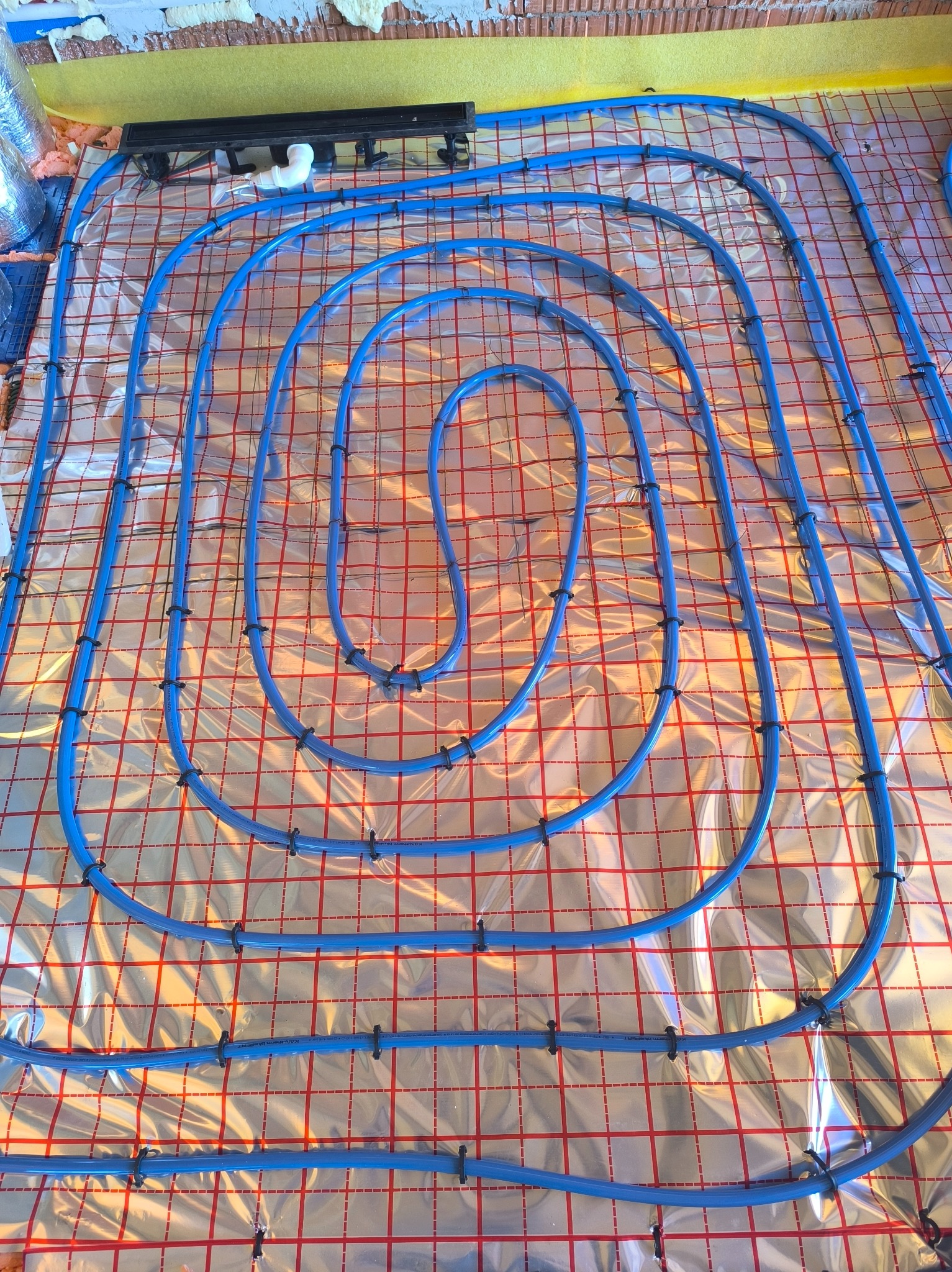 Błękitne rury ogrzewania podłogowego ułożone spiralnie na srebrnej folii z czerwoną siatką, przymocowane czarnymi klipsami. Widoczne fragmenty ścian.