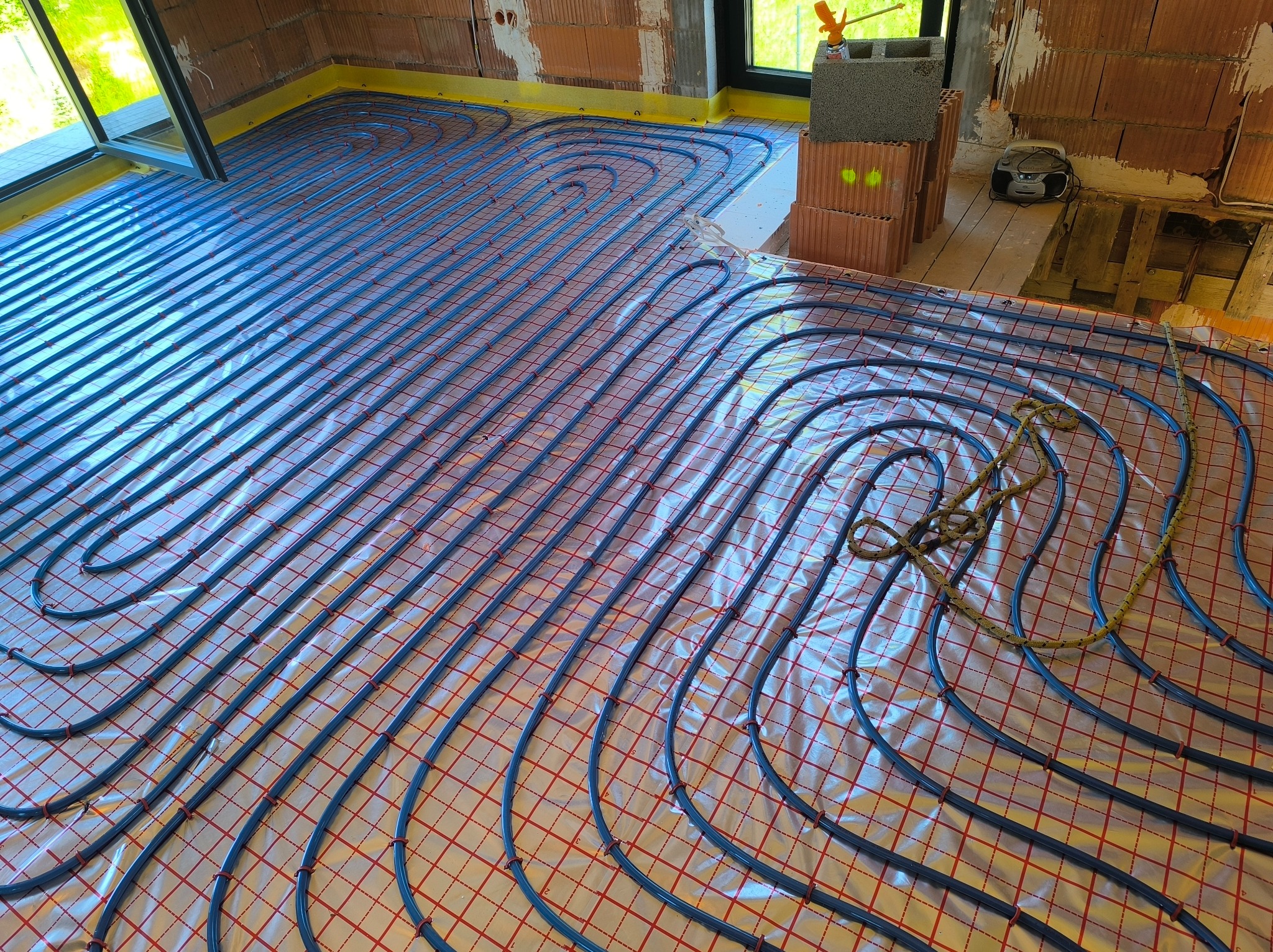 Instalacja ogrzewania podłogowego w Sadkowicach. Rury grzewcze ułożone na folii z siatką, widoczne cegły i okno. Nowoczesne rozwiązanie grzewcze.