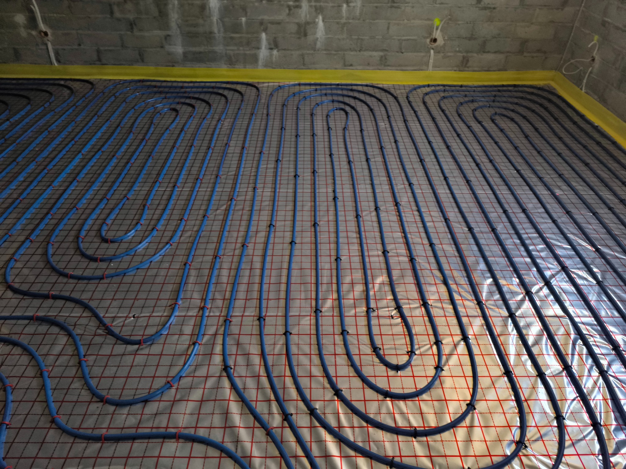Niebieskie rury ogrzewania podłogowego ułożone na folii z siatką, przymocowane czarnymi klipsami. Widoczna surowa ściana z betonu komórkowego.