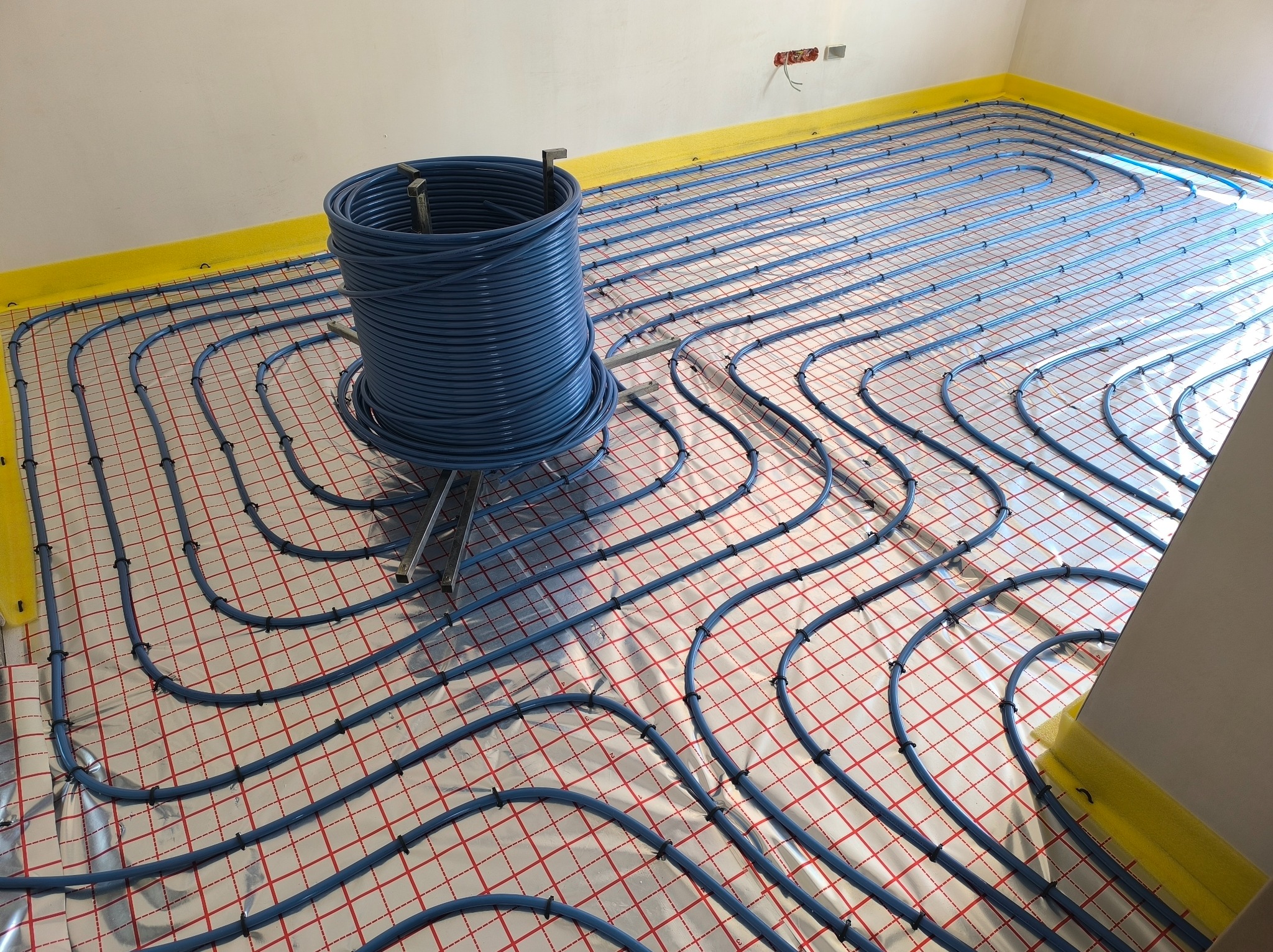 Niebieskie rury ogrzewania podłogowego ułożone na folii z czerwoną siatką, zwinięte w krąg na metalowym stojaku. Żółta taśma zabezpiecza ściany.