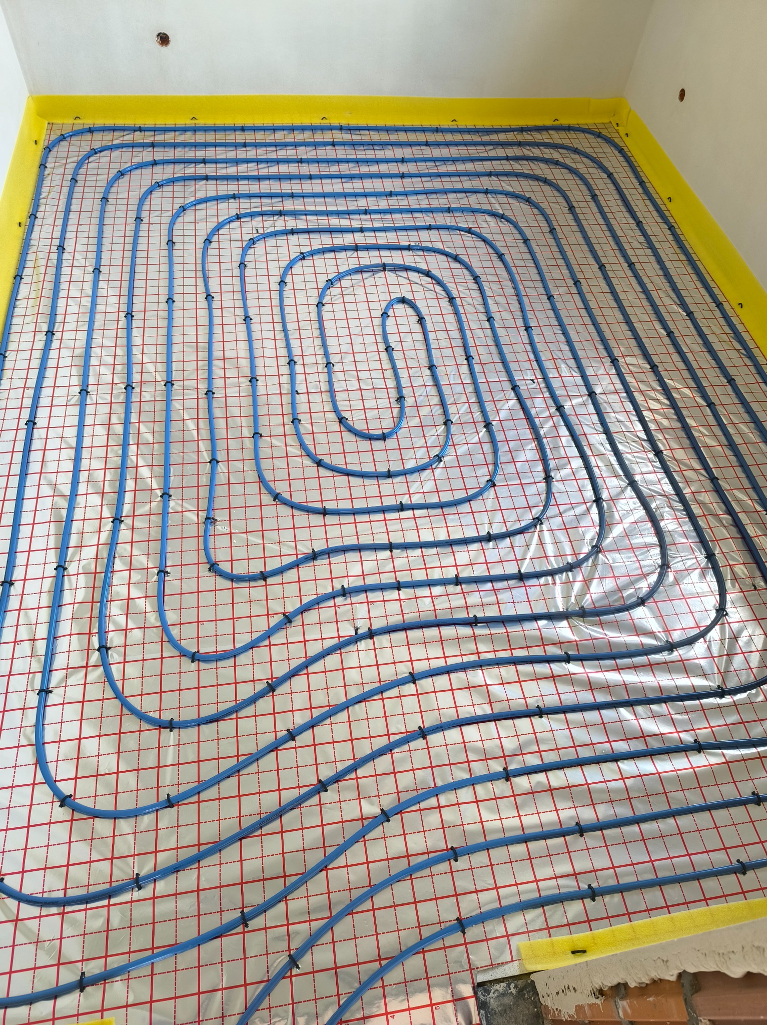 Niebieskie rury ogrzewania podłogowego ułożone spiralnie na folii z czerwoną kratką, przymocowane czarnymi klipsami, z żółtą taśmą brzegową.