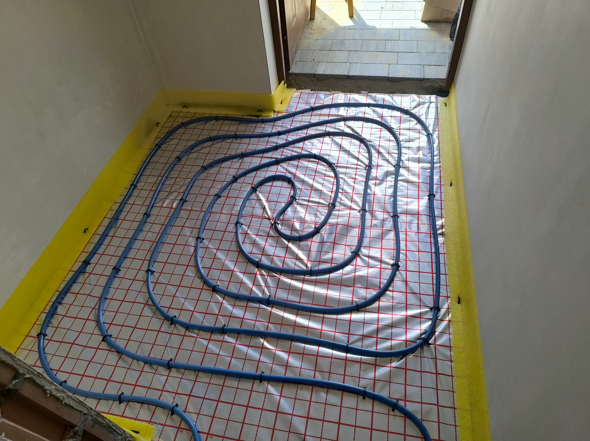 Niebieskie rury ogrzewania podłogowego ułożone spiralnie na folii z czerwoną siatką, przytwierdzone czarnymi uchwytami, z żółtą pianką izolacyjną przy ścianach.