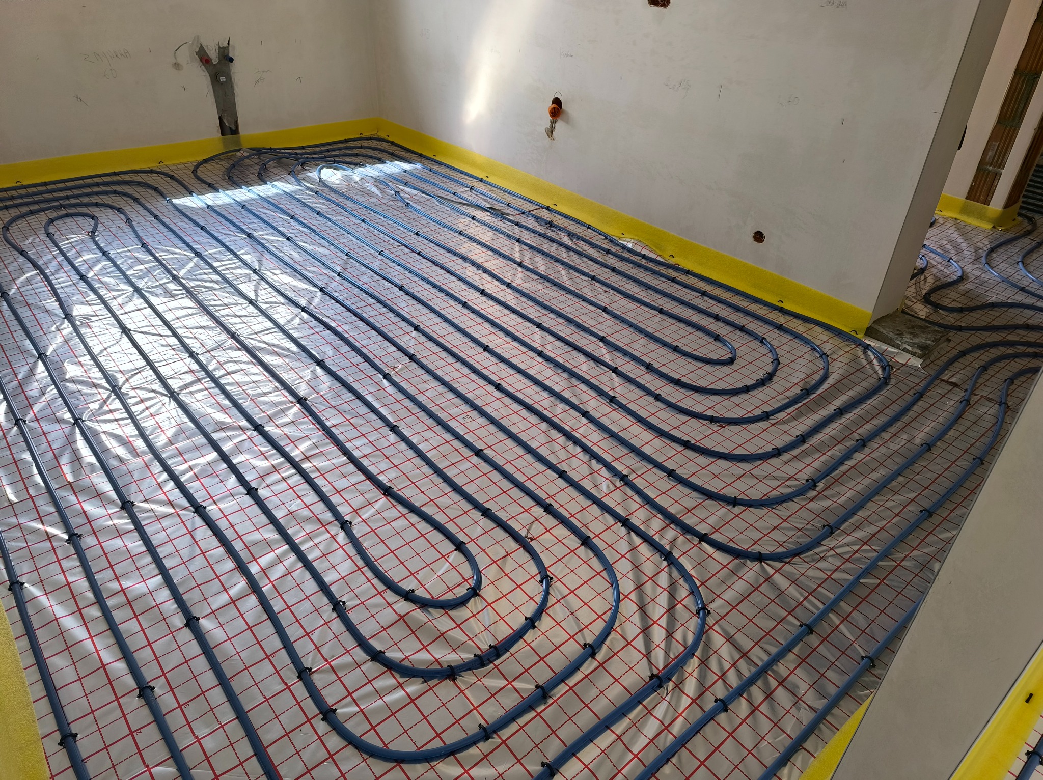 Instalacja ogrzewania podłogowego: niebieskie rury ułożone na folii z siatką w pomieszczeniu w trakcie remontu. Żółta taśma brzegowa wzdłuż ścian.