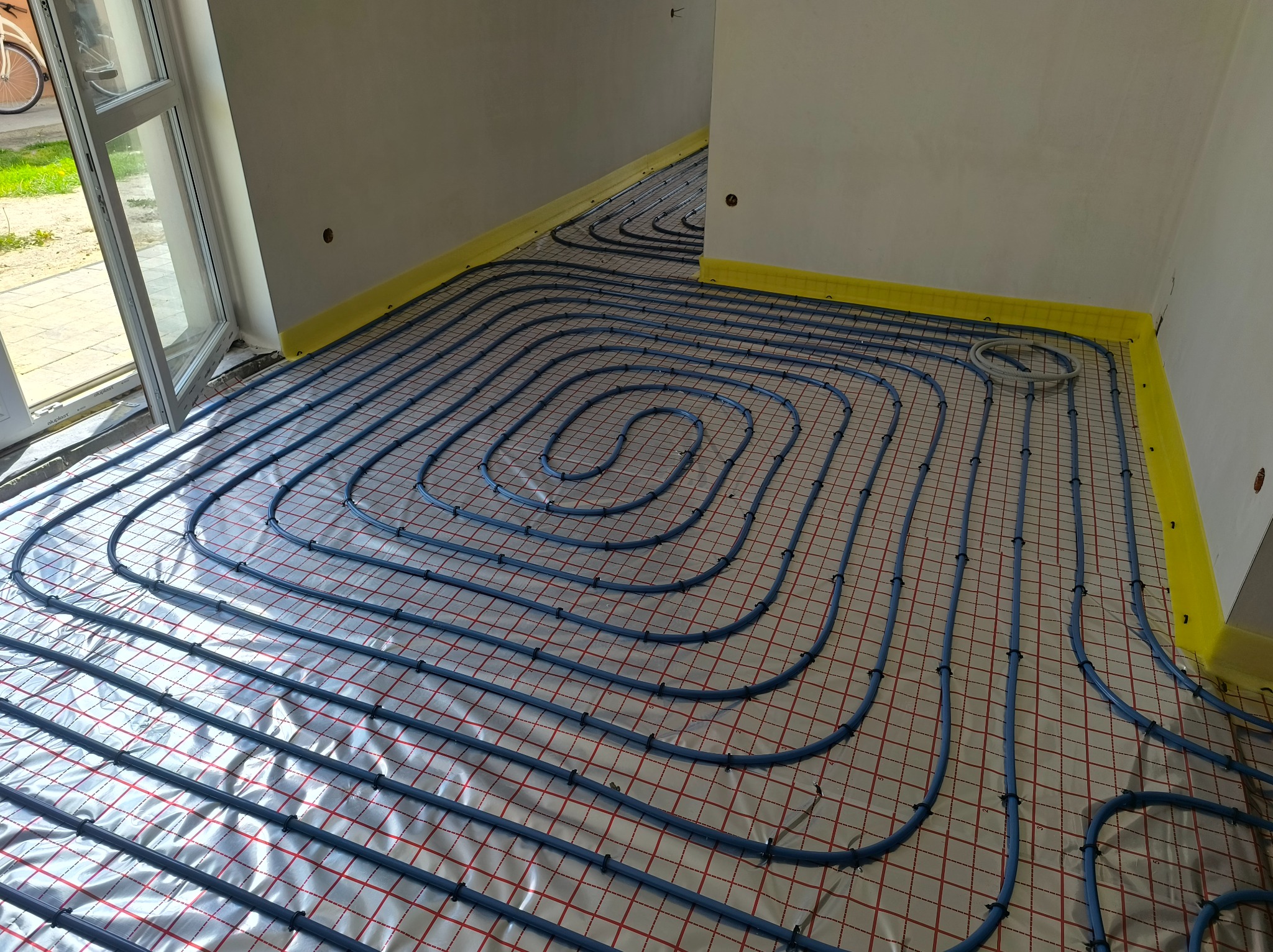 Niebieskie rury ogrzewania podłogowego, ułożone na folii z siatką, widoczne żółte paski izolacji przy ścianach i otwarte okno w jasnym wnętrzu.