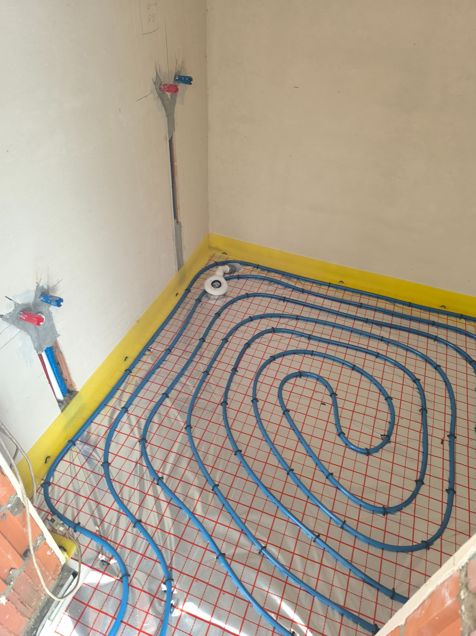 Niebieskie rury ogrzewania podłogowego ułożone na folii z siatką, widoczne przyłącza do instalacji w ścianie. Żółta taśma brzegowa.