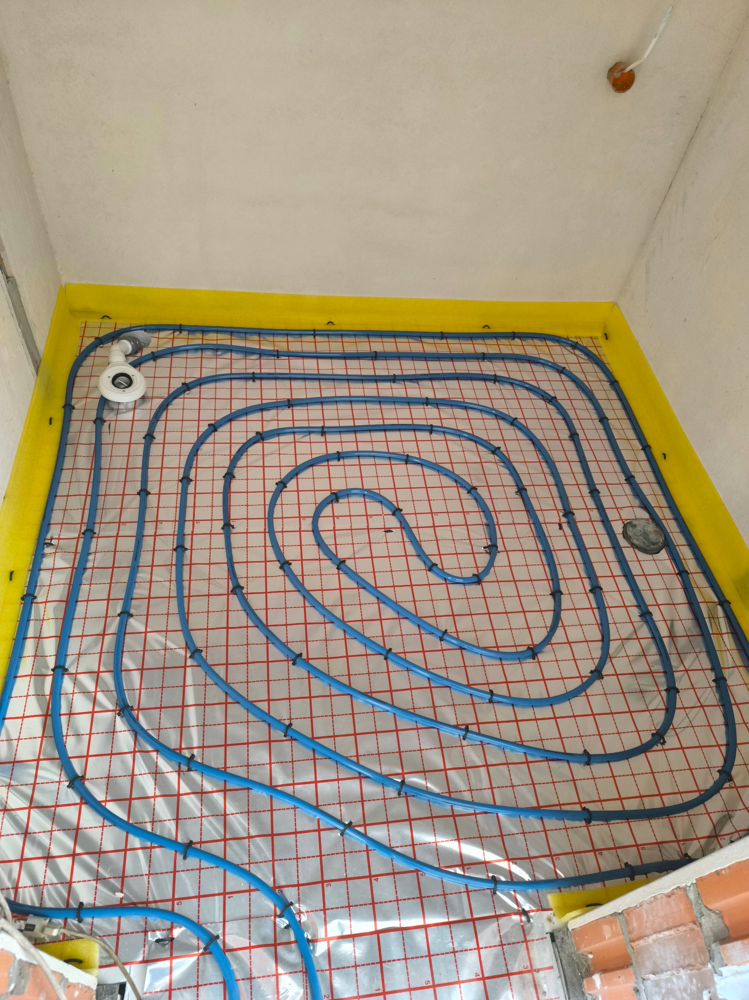 Niebieskie rury ogrzewania podłogowego ułożone spiralnie na izolacji z żółtą taśmą brzegową, widok z góry w surowym wnętrzu.