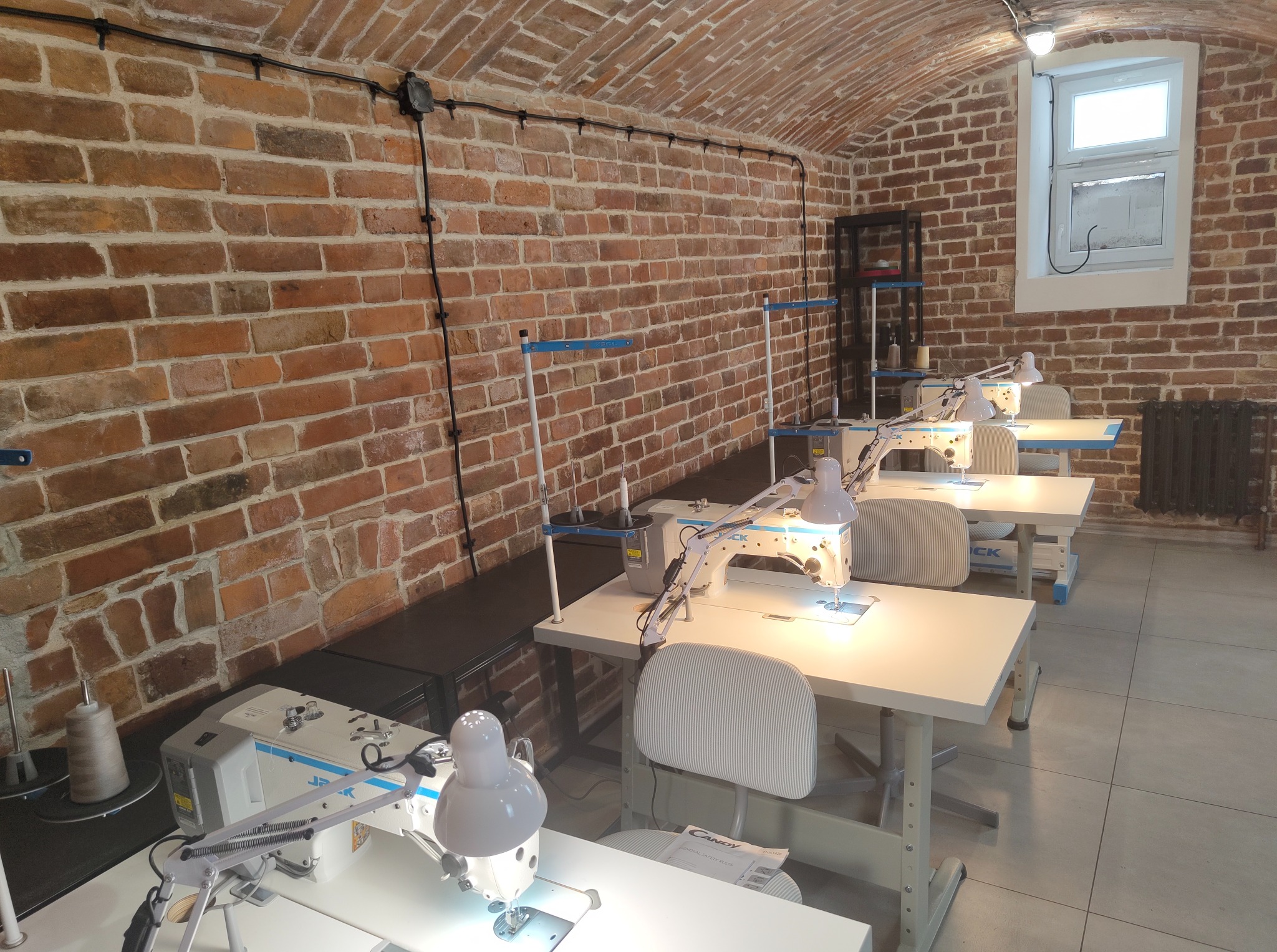 Wnętrze szwalni w Warszawie z rzędem białych maszyn do szycia marki Jack, ustawionych na stołach pod ceglanym, sklepionym sufitem.