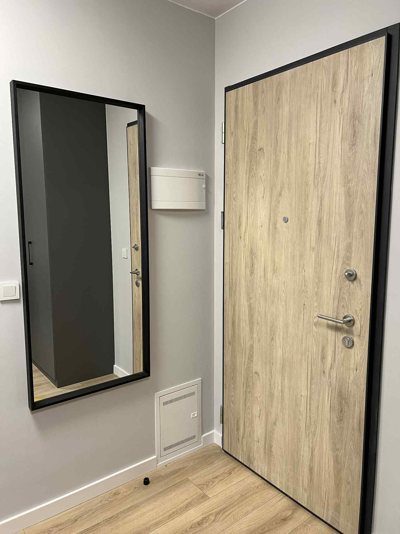 Nowoczesne drzwi wejściowe z drewnopodobnym fornirem i czarną ramą, oraz lustro w czarnej ramie w przedpokoju. Widoczne elementy wykończenia podłogi i ściany.