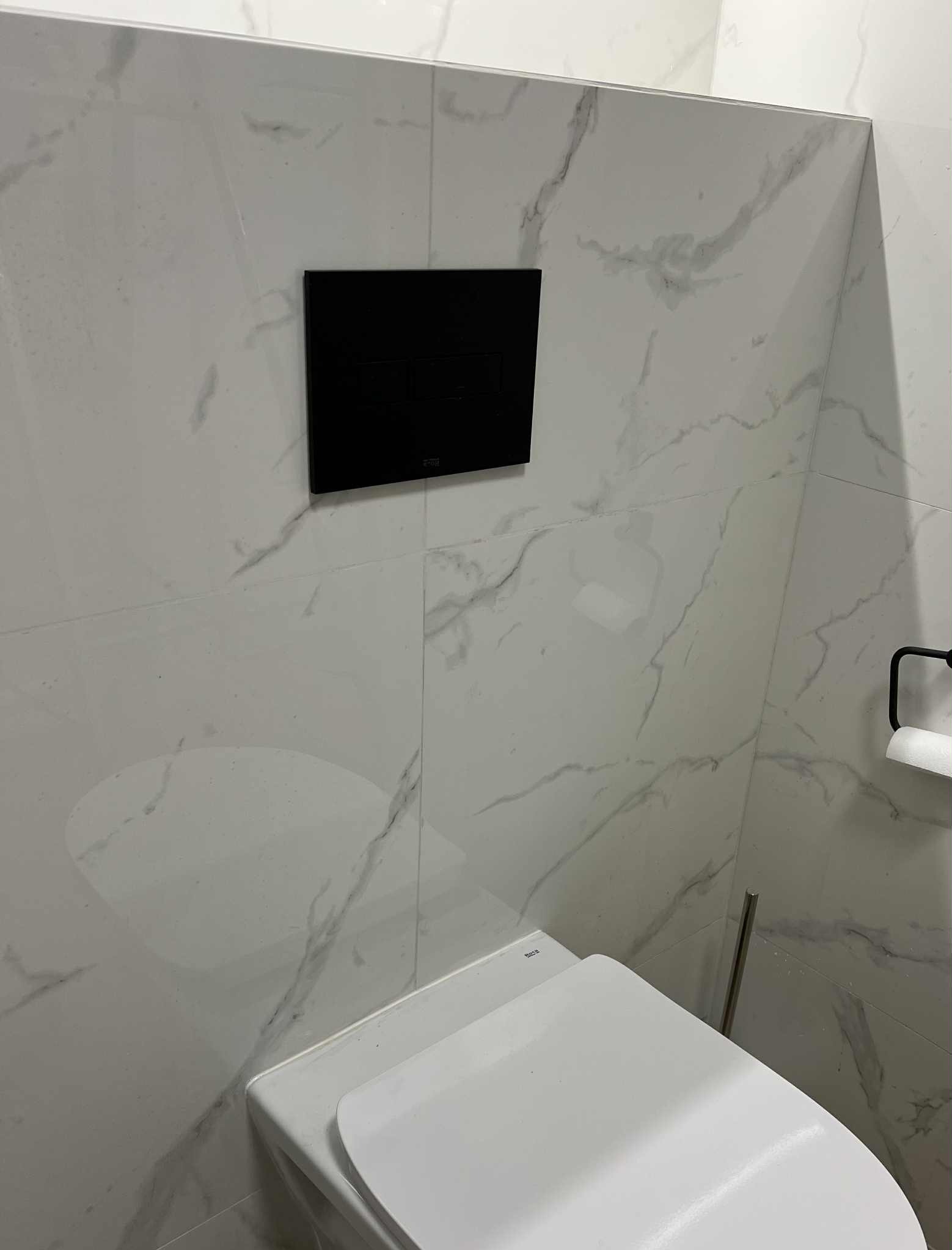 Wykończona łazienka: białe płytki imitujące marmur, wisząca toaleta z czarnym panelem sterującym. Minimalistyczny design, czystość i nowoczesność.