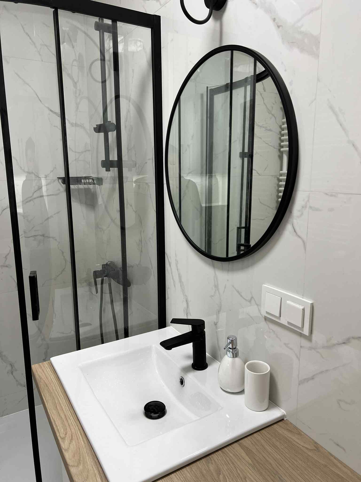 Nowoczesna łazienka: kabina prysznicowa z czarnymi ramami, okrągłe lustro, biała umywalka z drewnianym blatem i armaturą w czerni. Marmurowe płytki na ścianach.