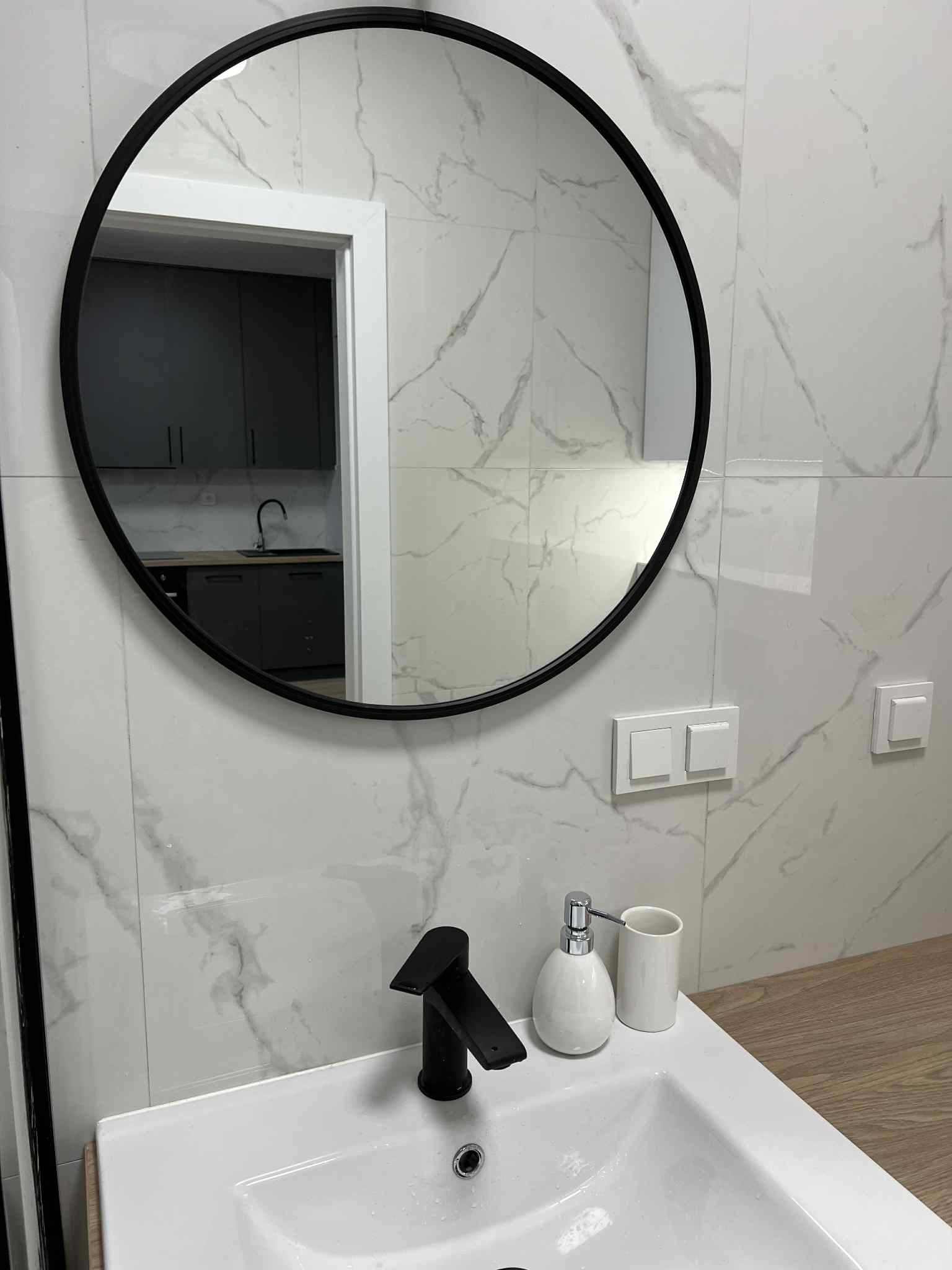 Wykończona łazienka: białe płytki imitujące marmur, okrągłe lustro z czarną ramą, czarna bateria umywalkowa i elementy dekoracyjne. Widok na aneks kuchenny.