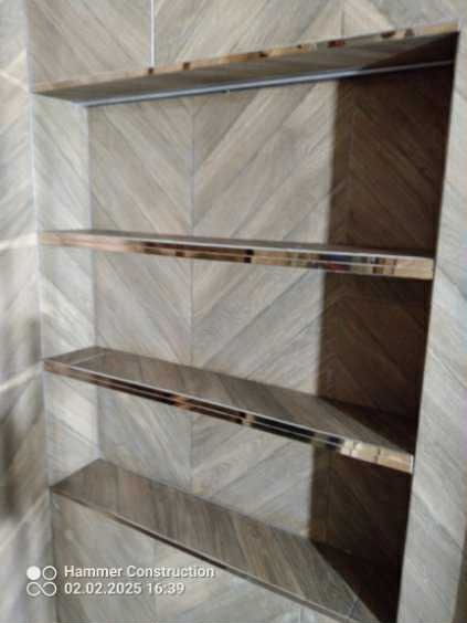 Narożne, podświetlone półki z płytek imitujących drewno, wbudowane we wnękę ściany wyłożonej płytkami w geometryczny wzór jodełki. Nowoczesny detal wykończenia łazienki.