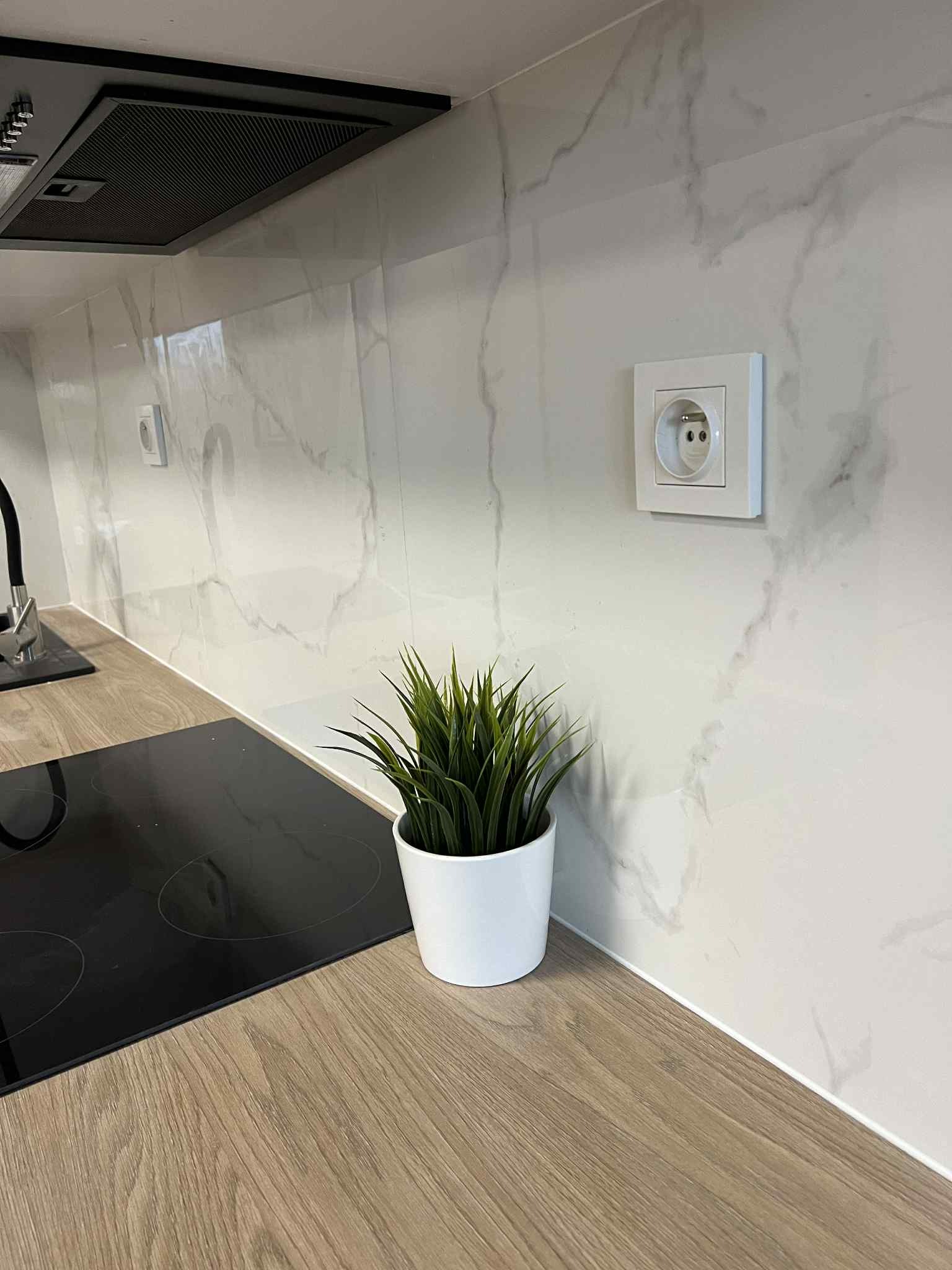 Biała marmurowa ściana nad blatem z płyty imitującej drewno, gniazdko elektryczne i czarny okap kuchenny. Na blacie doniczka z trawą.
