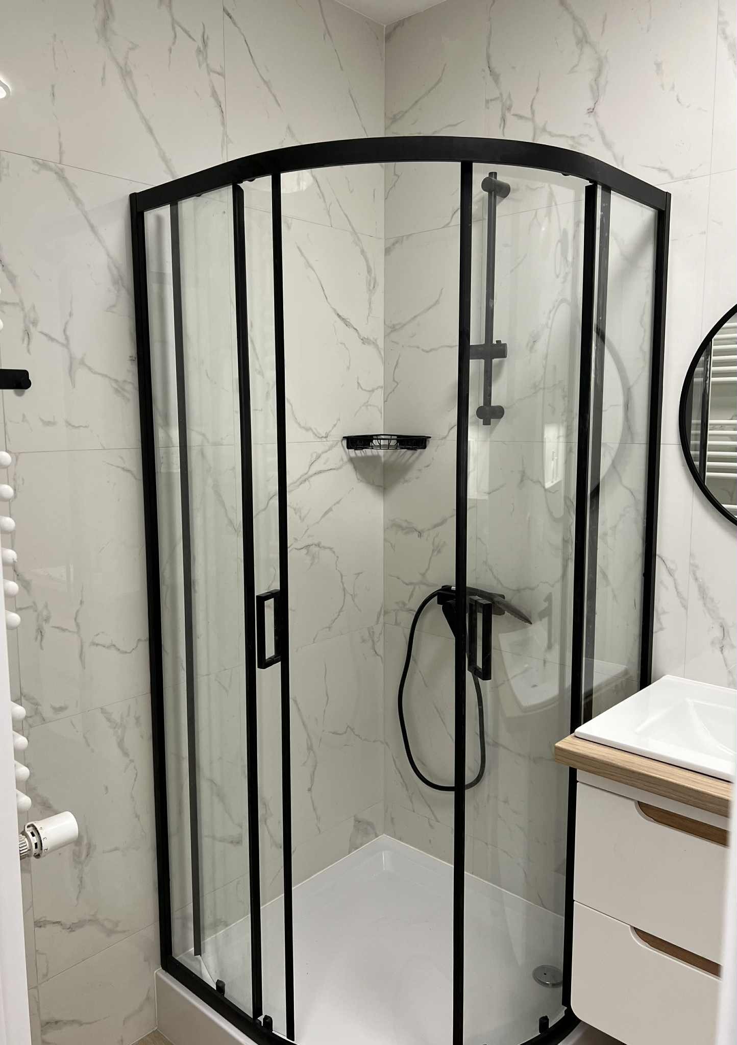 Kabina prysznicowa z czarną ramą na tle marmurowych płytek. Widoczna umywalka z szafką i okrągłe lustro. Nowoczesna aranżacja łazienki po remoncie.