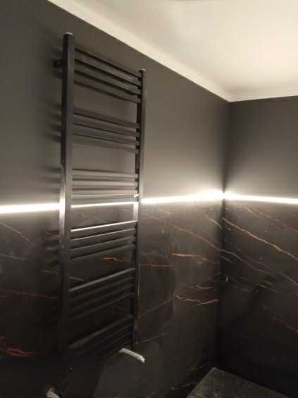 Czarny grzejnik drabinkowy na ścianie z ciemnego marmuru w łazience, oświetlonej subtelnym światłem LED, tworząc nowoczesny i elegancki wygląd.