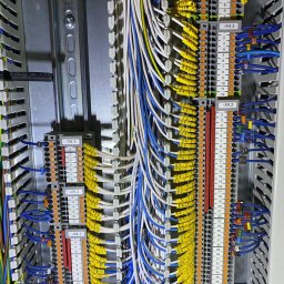 EKSS Jakub Klimontowicz - Rozdzielnia elektryczna z precyzyjnie ułożonymi kablami w kolorach: białym, niebieskim i żółtym z oznaczeniami. Widoczne listwy zaciskowe i metalowa szyna DIN.