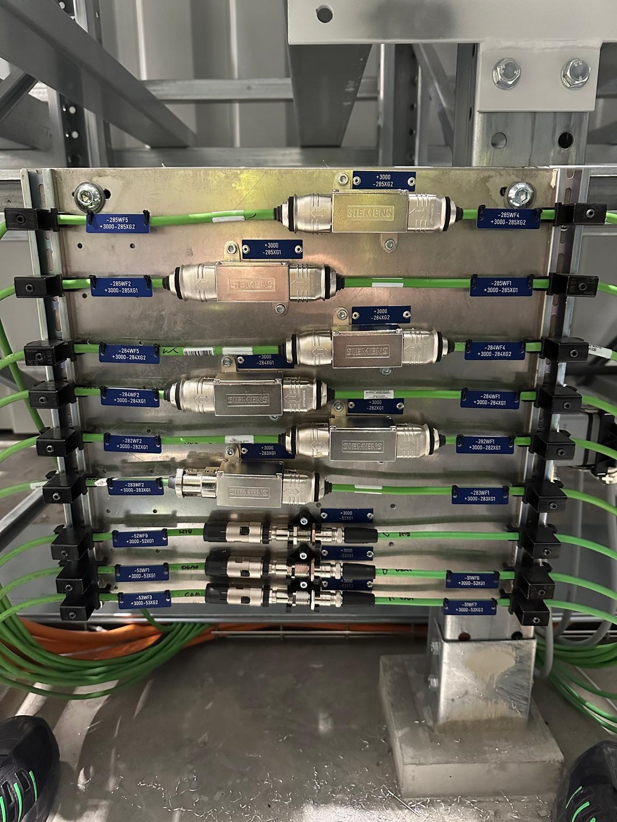 Szczegółowe ujęcie panelu z przewodami i złączkami Siemens, oznaczonymi etykietami, zamontowanego w metalowej konstrukcji. Widoczne kable w kolorze zielonym i pomarańczowym.