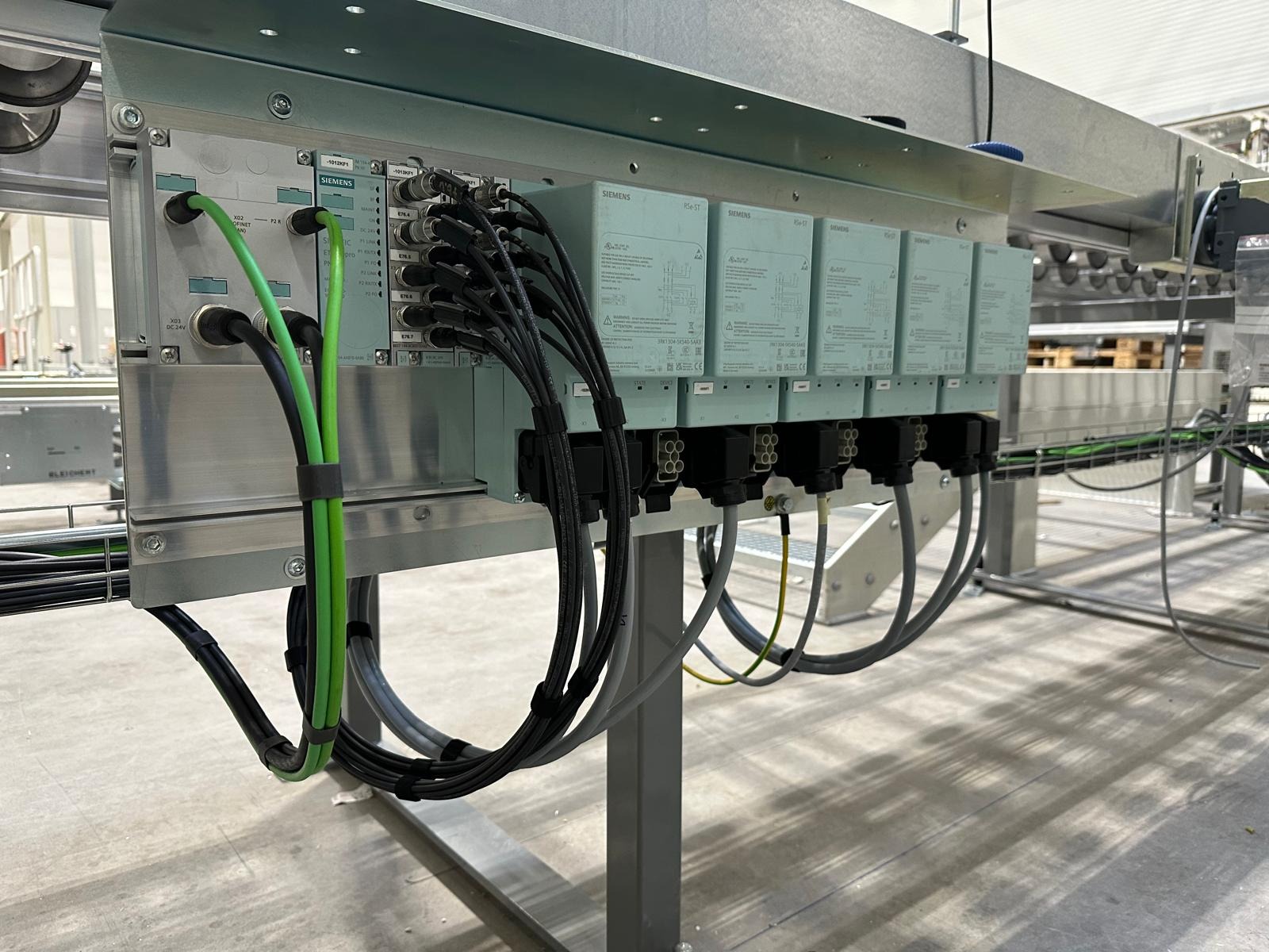 Panel sterowania Siemens z licznymi kablami i złączami, zamontowany na metalowej konstrukcji. Widoczne moduły sterujące i okablowanie w kolorach czarnym, szarym i zielonym.