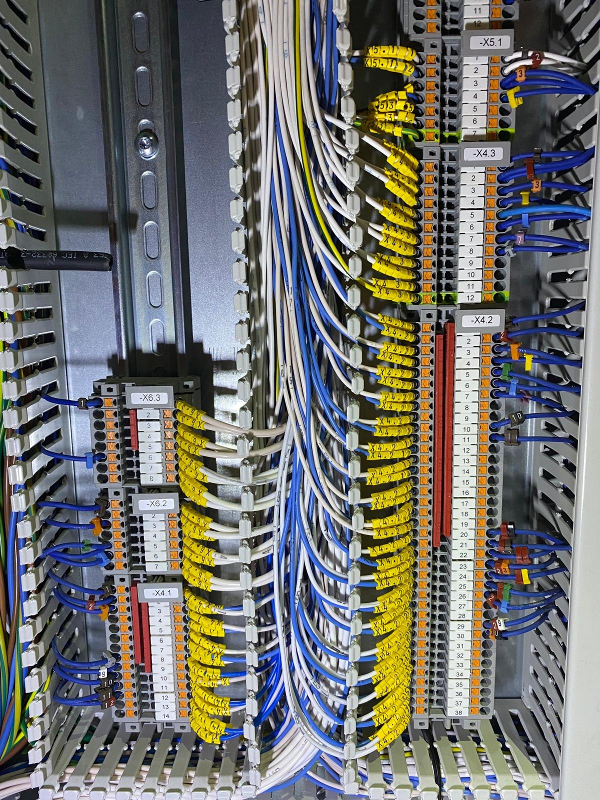 Rozdzielnia elektryczna z precyzyjnie ułożonymi kablami w kolorach: białym, niebieskim i żółtym z oznaczeniami. Widoczne listwy zaciskowe i metalowa szyna DIN.