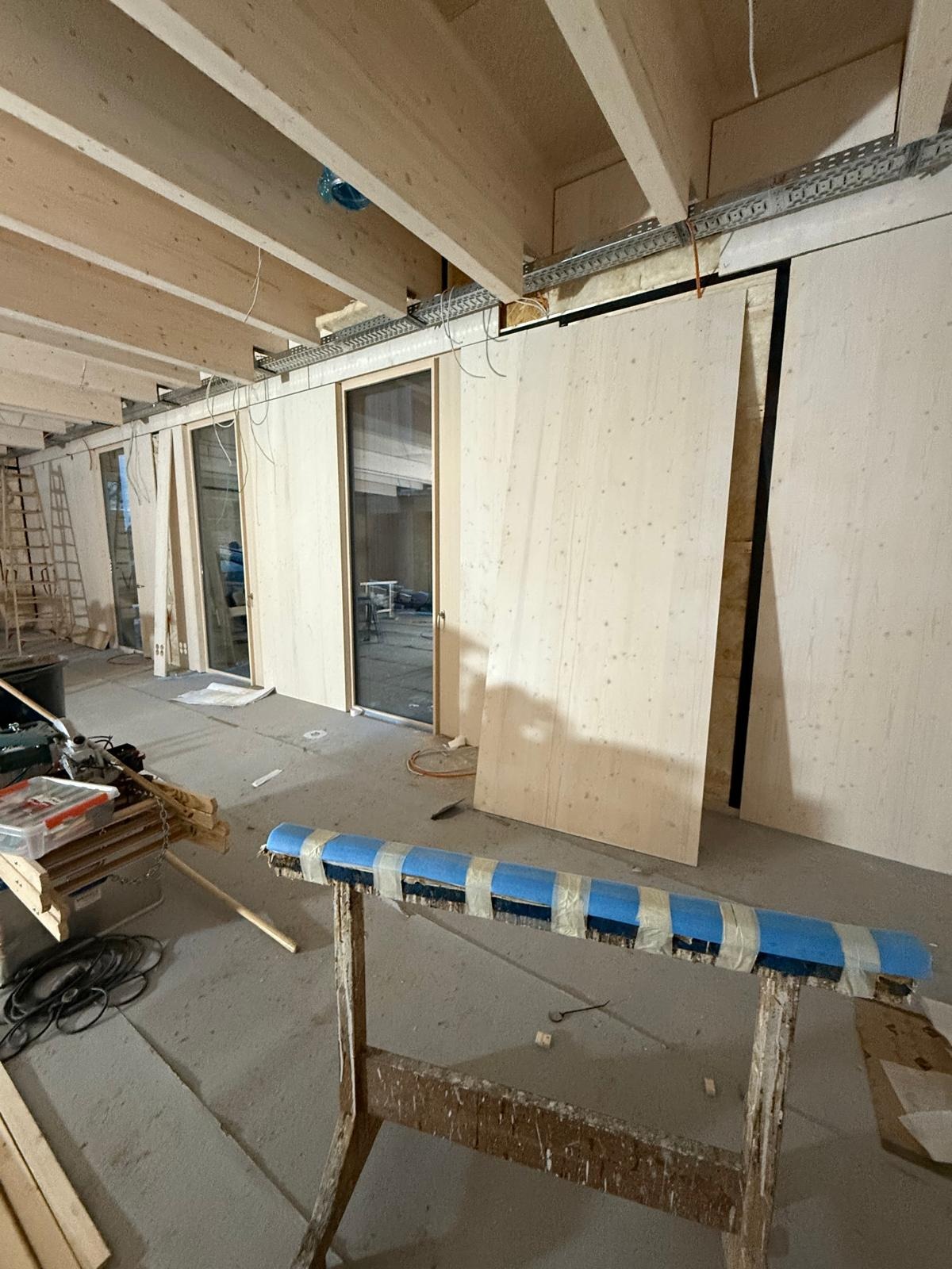 Wnętrze w trakcie prac wykończeniowych. Widoczne drewniane elementy konstrukcyjne, ściany z płyt oraz narzędzia na pierwszym planie. Surowy stan, instalacje w trakcie montażu.