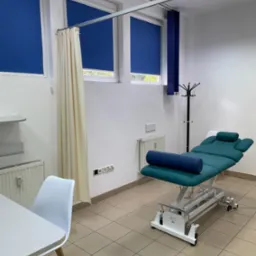 Jasne wnętrze gabinetu z profesjonalnym, regulowanym kozetką rehabilitacyjną w kolorze turkusowym i niebieskim wałkiem. Okna z roletami w odcieniach niebieskiego.