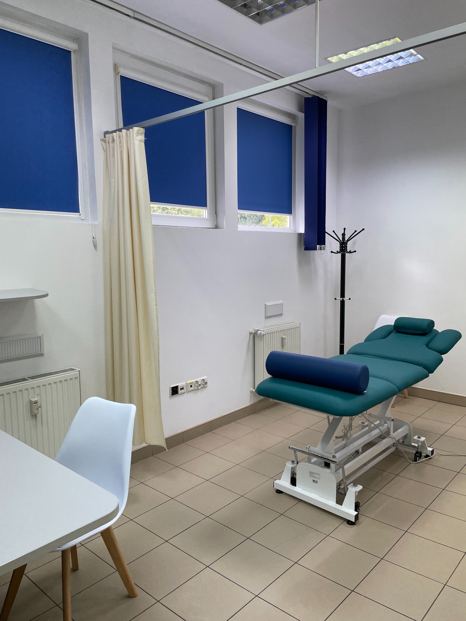 Jasne wnętrze gabinetu z profesjonalnym, regulowanym kozetką rehabilitacyjną w kolorze turkusowym i niebieskim wałkiem. Okna z roletami w odcieniach niebieskiego.