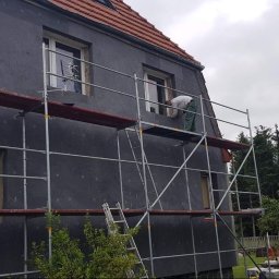 TENRAI SPÓŁKA Z OGRANICZONĄ ODPOWIEDZIALNOŚCIĄ - Dom w trakcie ocieplania styropianem, widoczne rusztowania i pracownik przy oknie. Część elewacji jest już ocieplona, a część jeszcze nie.