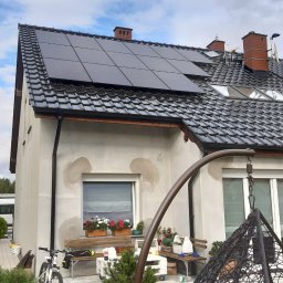 TENRAI SPÓŁKA Z OGRANICZONĄ ODPOWIEDZIALNOŚCIĄ - Dom z panelami fotowoltaicznymi na dachu z czarnej dachówki. Widoczne łaty po naprawach elewacji, rower i huśtawka wisząca w ogrodzie. Słoneczny dzień.