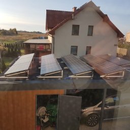 TENRAI SPÓŁKA Z OGRANICZONĄ ODPOWIEDZIALNOŚCIĄ - Instalacja fotowoltaiczna na dachu garażu przylegającego do domu jednorodzinnego. Widok z okna, w tle zabudowa jednorodzinna i pola.