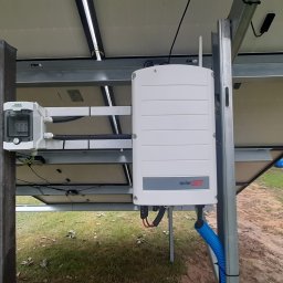 TENRAI SPÓŁKA Z OGRANICZONĄ ODPOWIEDZIALNOŚCIĄ - Widok od spodu na zamontowane panele fotowoltaiczne z widocznym inwerterem SolarEdge i skrzynką przyłączeniową na metalowej konstrukcji wsporczej.