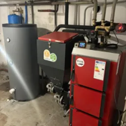 Nowoczesny, zainstalowany piec CO z podajnikiem i zasobnikiem na paliwo stałe, widoczne rury instalacji grzewczej. Kotłownia w piwnicy.