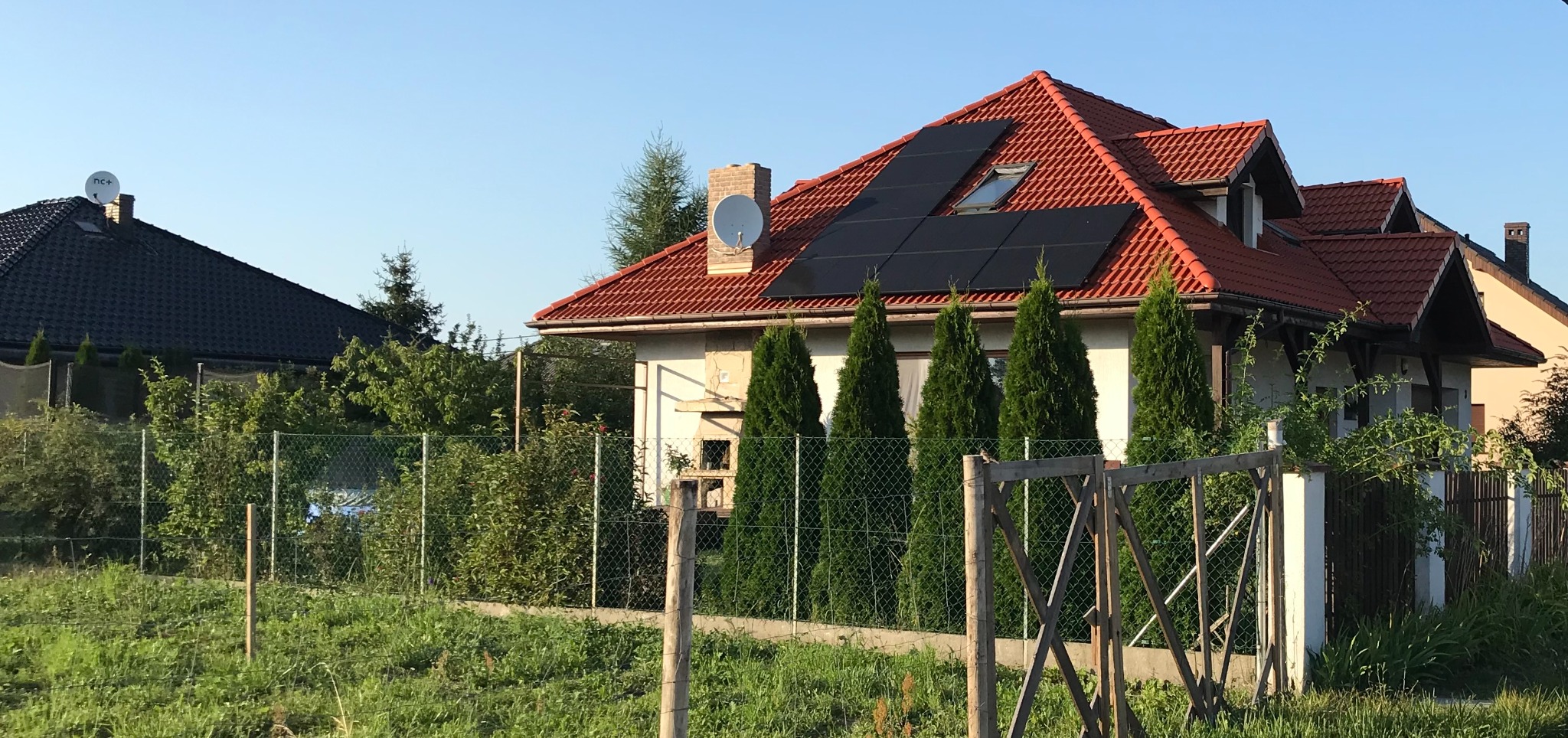 Dom jednorodzinny z panelami fotowoltaicznymi na czerwonym dachu, widoczny zza ogrodzenia. Na kominie antena satelitarna. Słoneczny dzień.