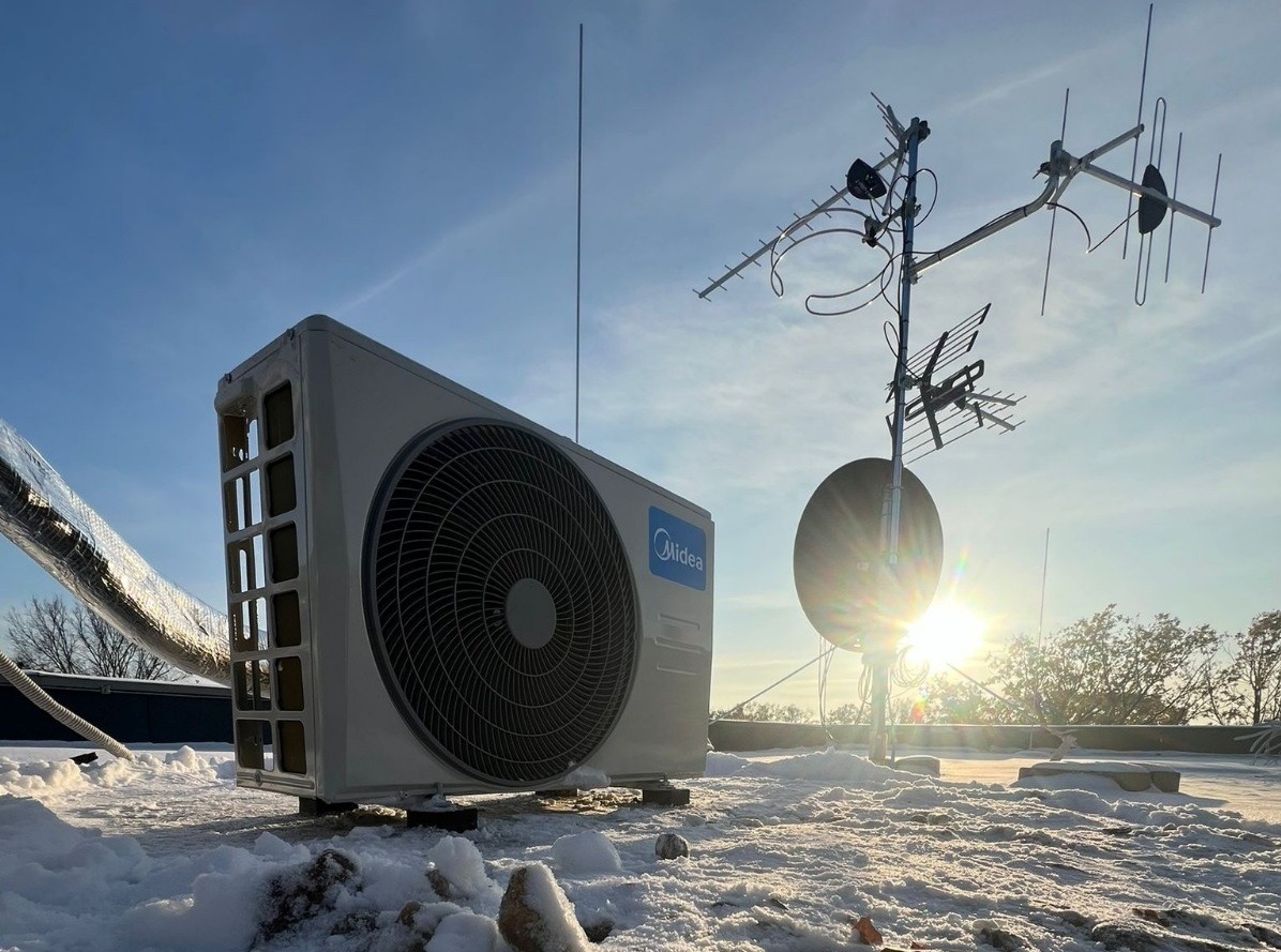 Biała jednostka zewnętrzna klimatyzacji Midea na zaśnieżonym dachu, z anteną satelitarną w tle i promieniami słońca. Widok z dołu, podkreślający instalację.