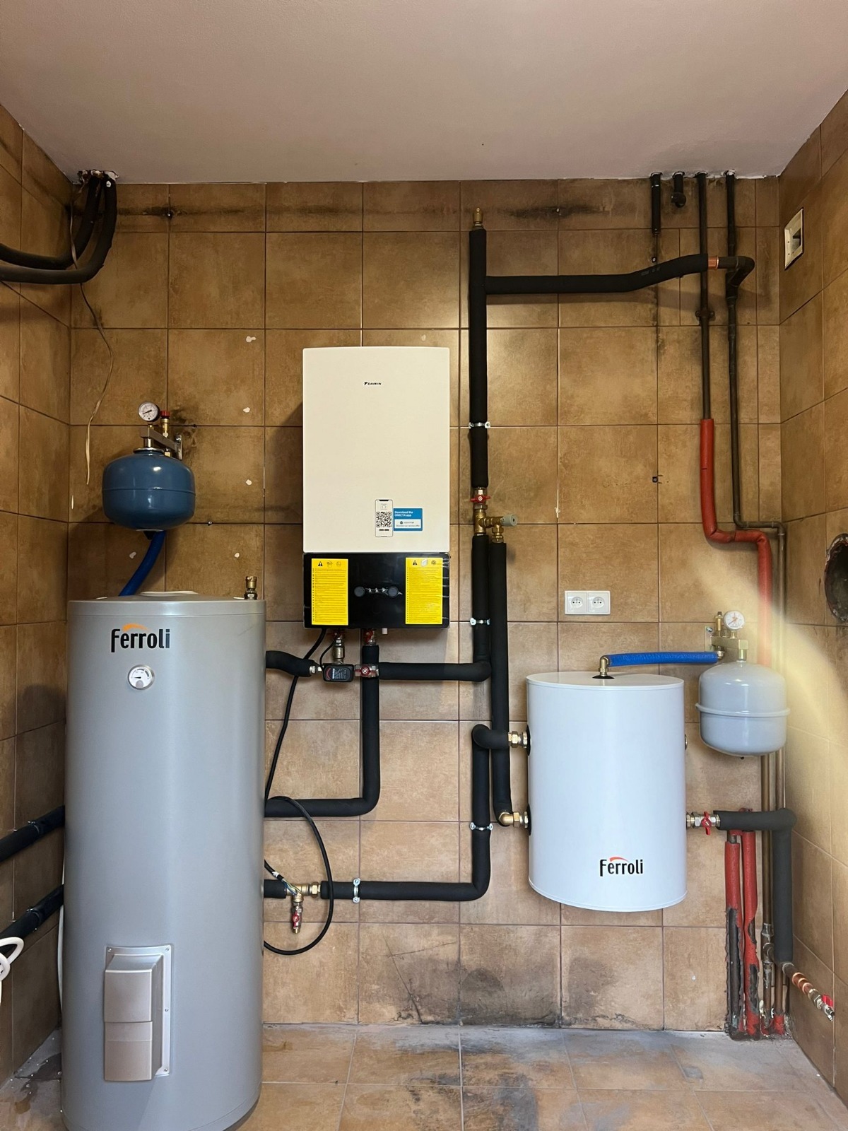 Zestaw urządzeń grzewczych Ferroli i Daikin z rurami w izolacji, zamontowany na tle brązowych płytek. Widoczne zbiorniki, pompa ciepła i elementy instalacji.