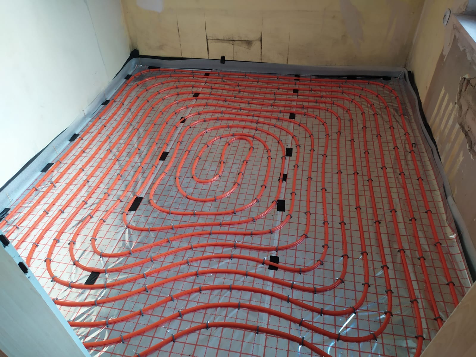 Pomarańczowe rury ogrzewania podłogowego ułożone na siatce w pomieszczeniu przed wylaniem posadzki, widoczne mocowania i folia izolacyjna przy ścianach.
