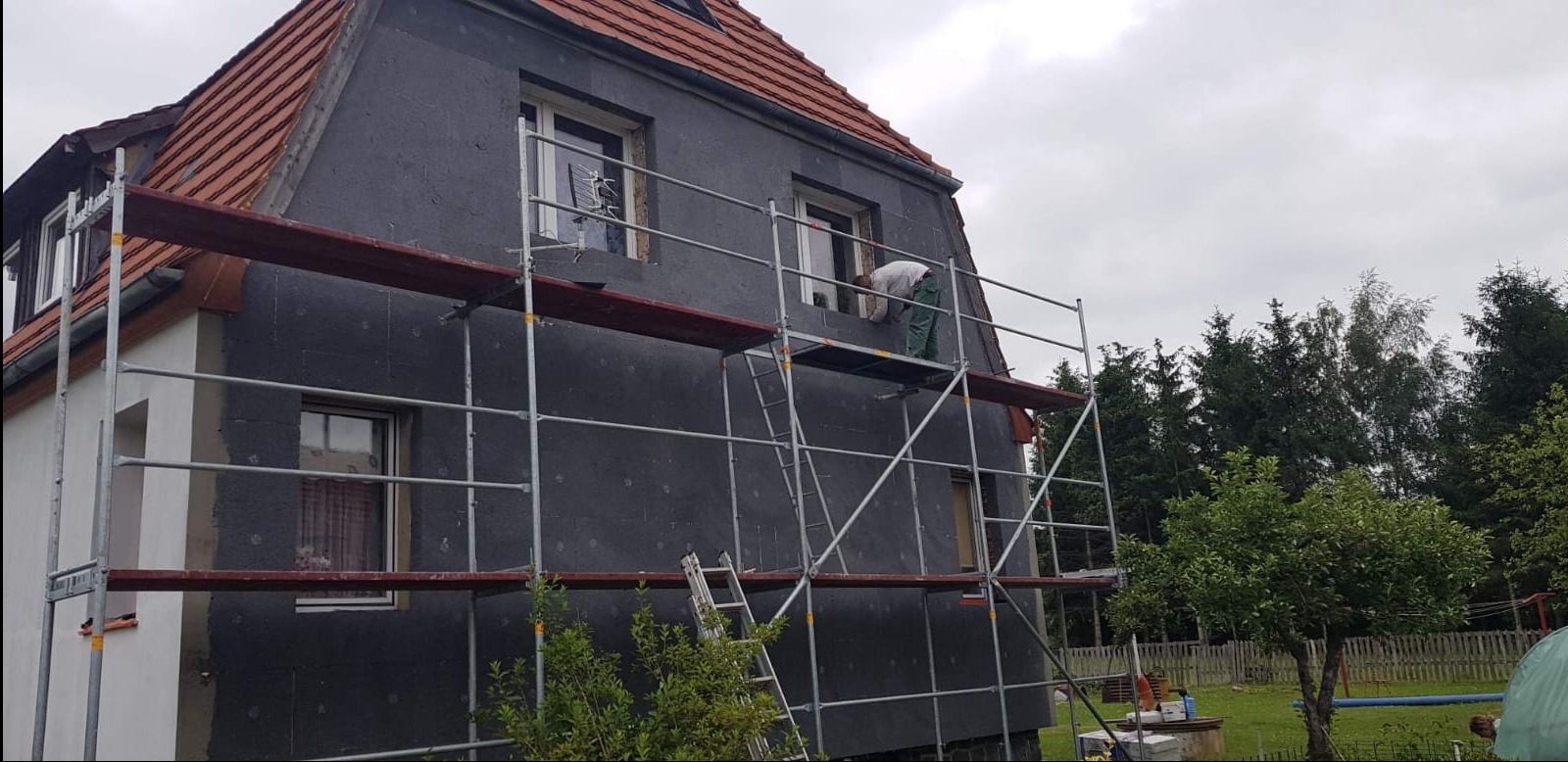 Dom w trakcie ocieplania styropianem, widoczne rusztowania i pracownik przy oknie. Część elewacji jest już ocieplona, a część jeszcze nie.