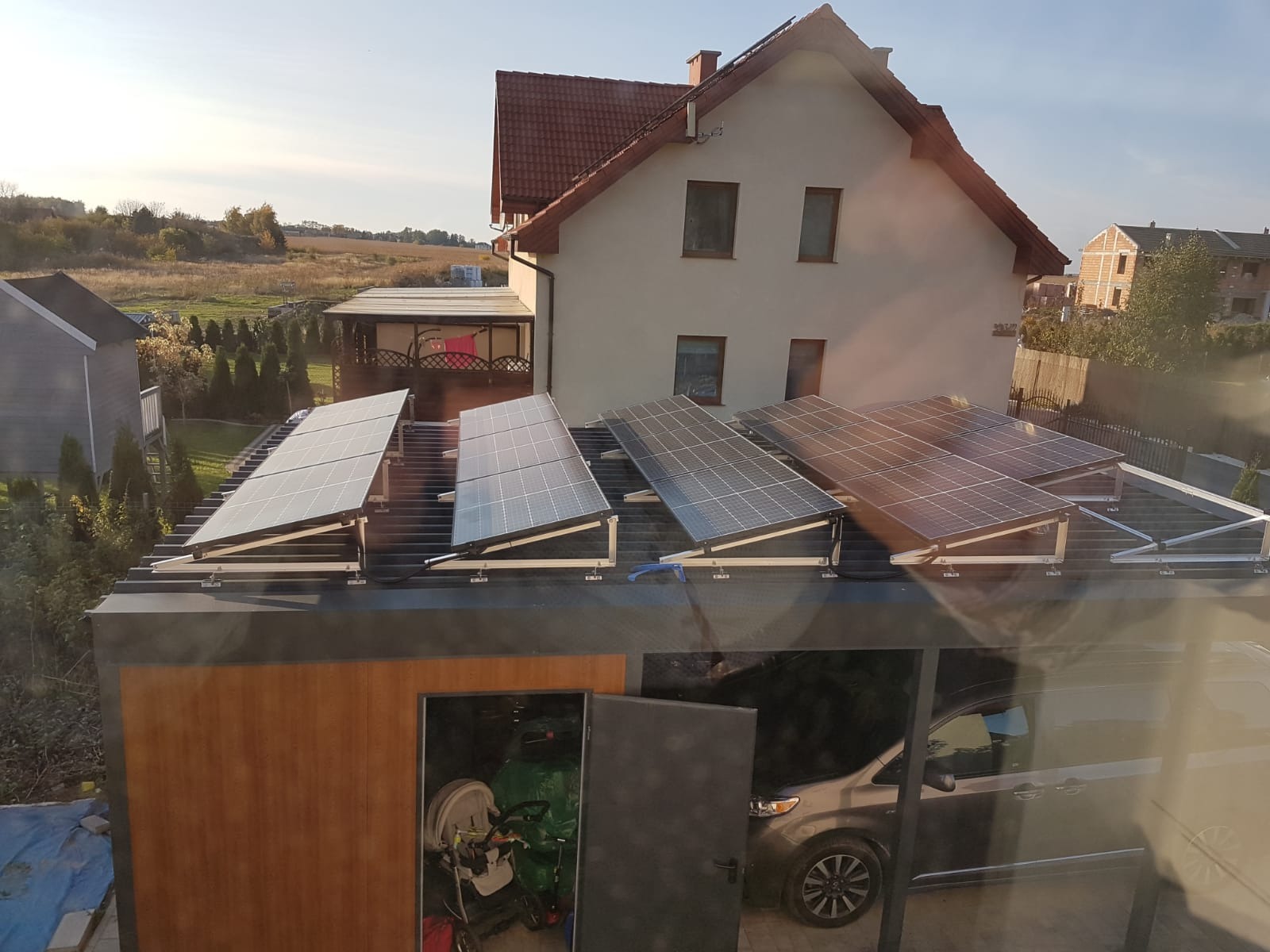 Instalacja fotowoltaiczna na dachu garażu przylegającego do domu jednorodzinnego. Widok z okna, w tle zabudowa jednorodzinna i pola.