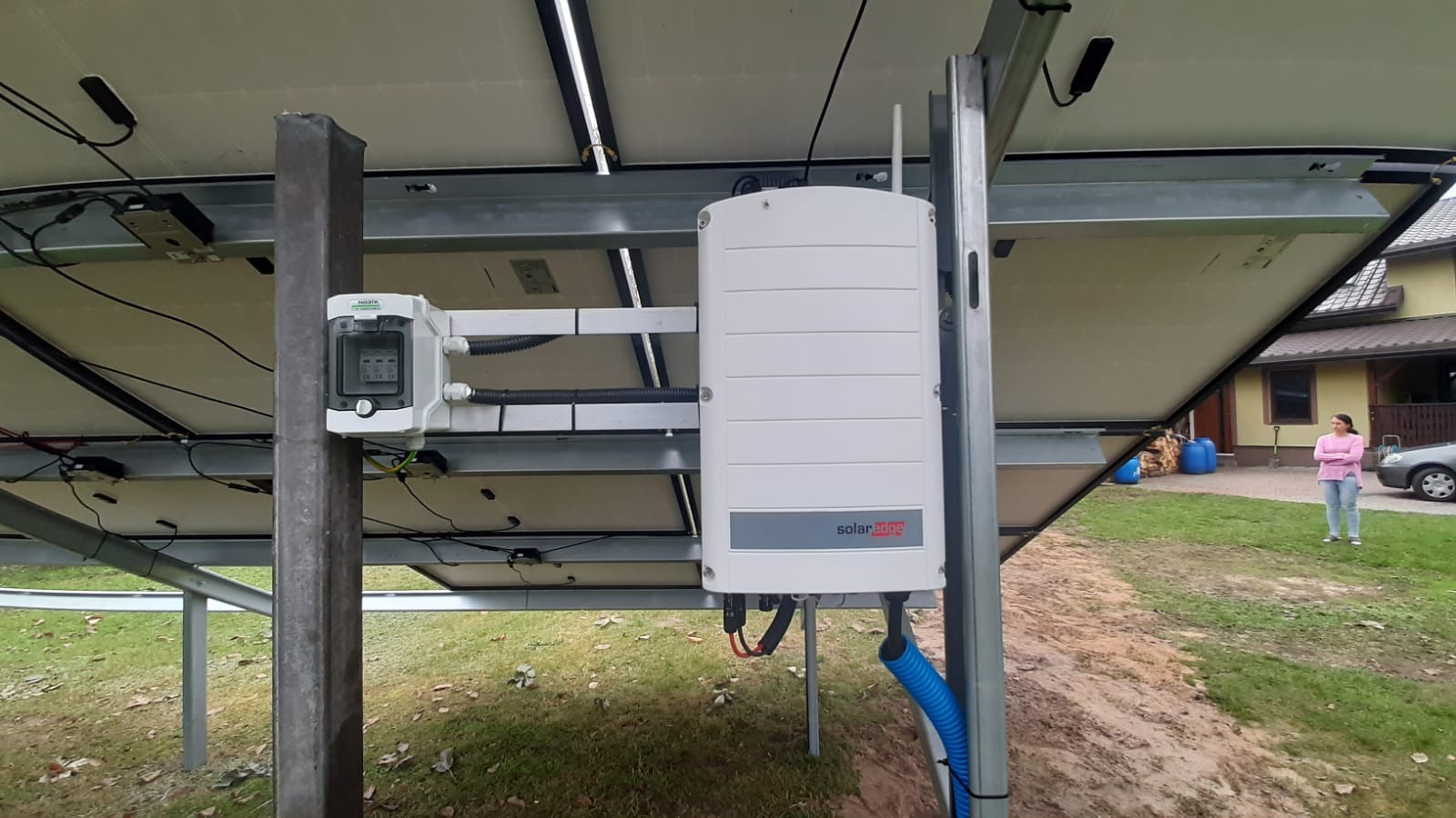 Widok od spodu na zamontowane panele fotowoltaiczne z widocznym inwerterem SolarEdge i skrzynką przyłączeniową na metalowej konstrukcji wsporczej.