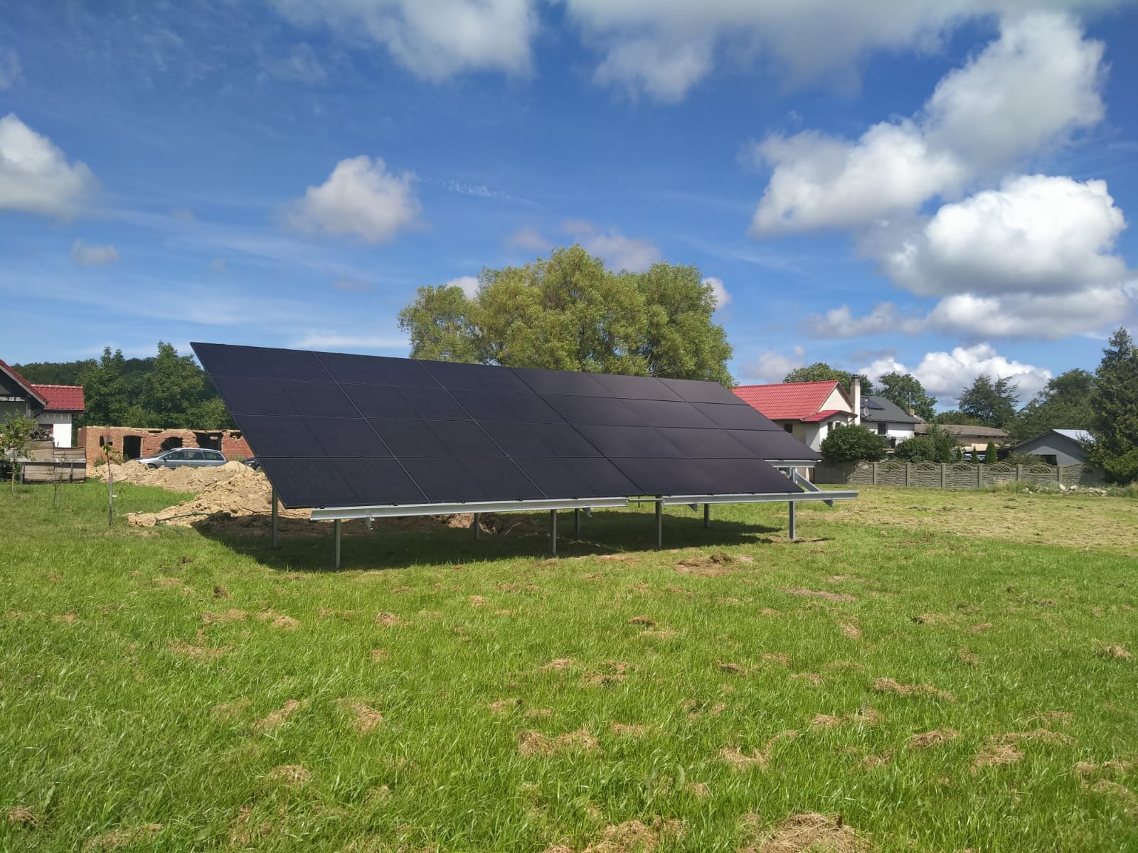 Czarny panel fotowoltaiczny na metalowej konstrukcji, umieszczony na trawiastym terenie z budynkami w tle, pod błękitnym niebem z chmurami.