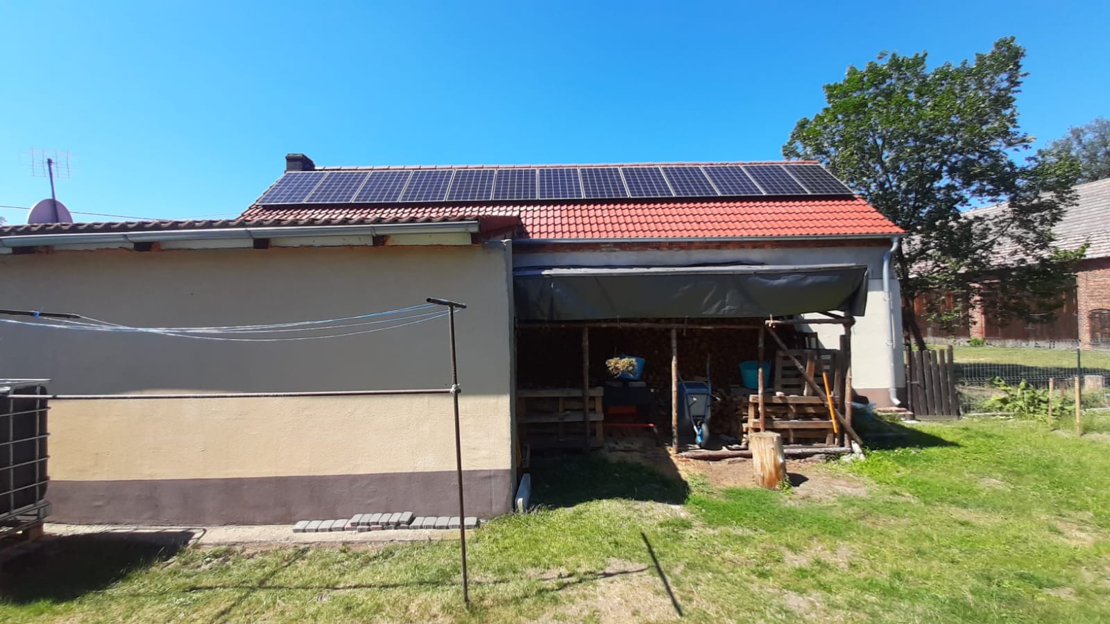 Panele słoneczne na dachu budynku gospodarczego z czerwoną dachówką, widoczne z poziomu trawnika w słoneczny dzień. Antena satelitarna na sąsiednim budynku.