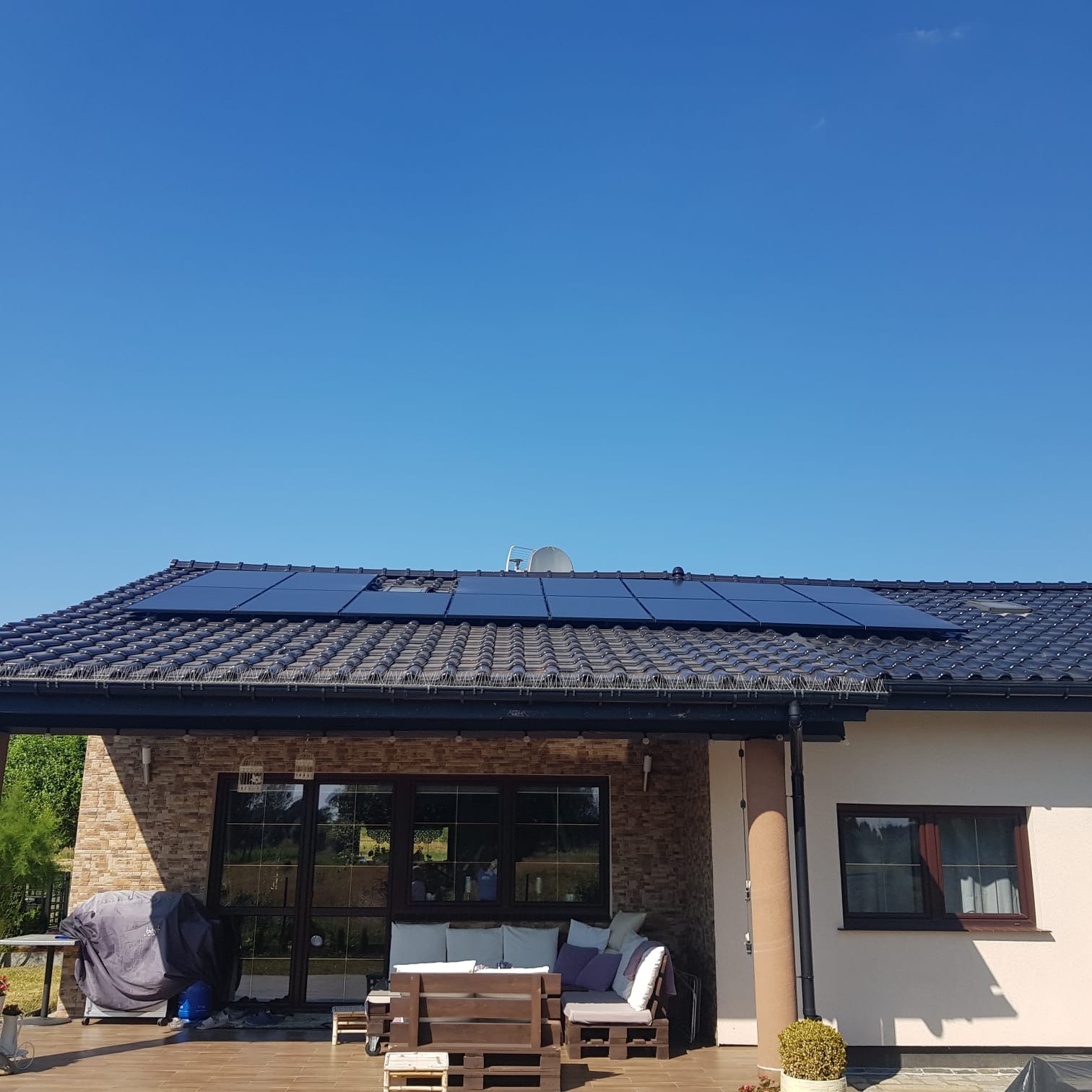Panele fotowoltaiczne na dachu domu jednorodzinnego z tarasem w słoneczny dzień. Widoczne czarne panele i brązowa dachówka. Na tarasie meble ogrodowe.