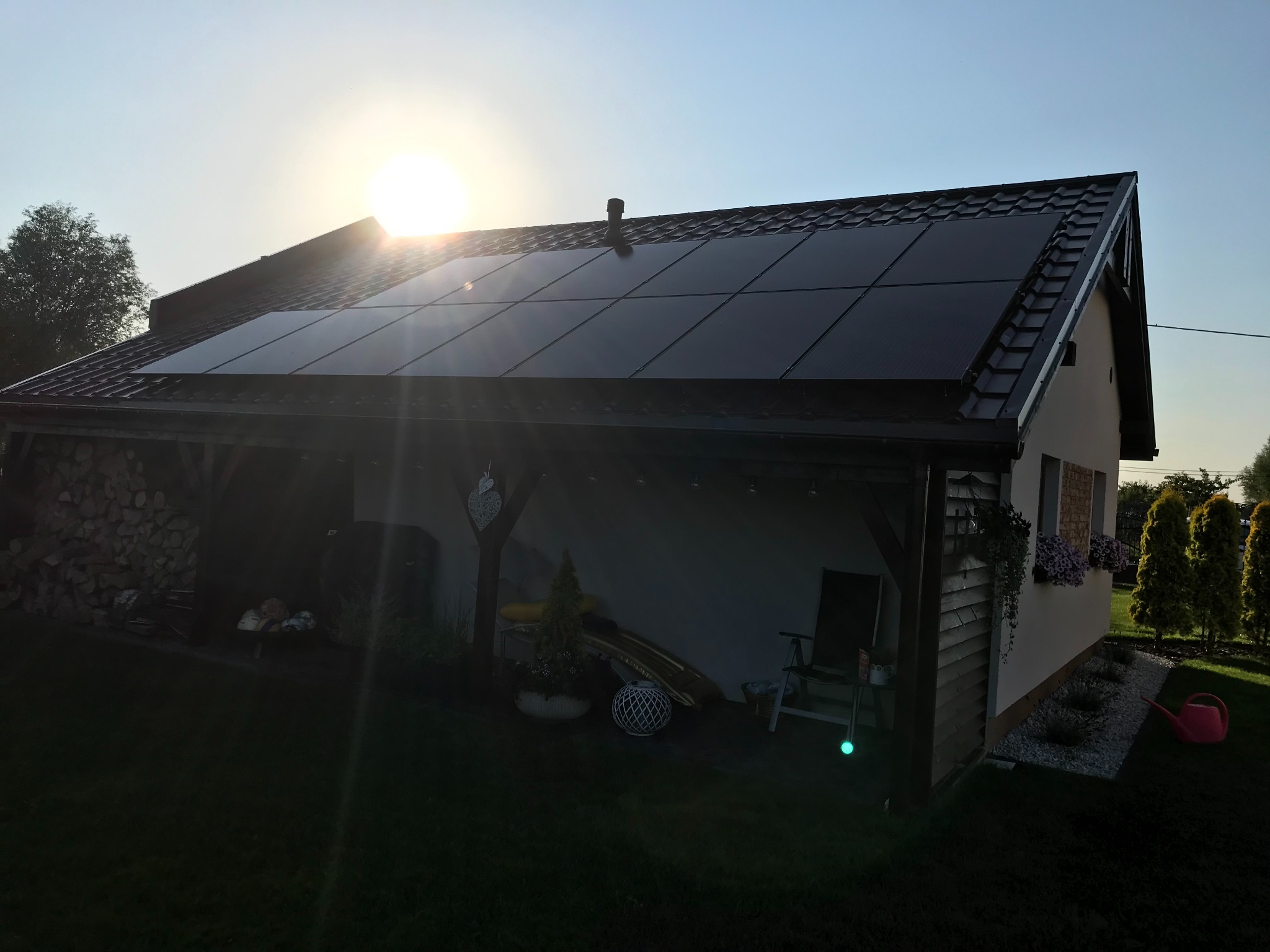 Dom z panelami fotowoltaicznymi na dachu, widok z ogrodu w słoneczny dzień. Na pierwszym planie trawnik, w tle budynek z drewnianą wiatą i ułożonym drewnem.