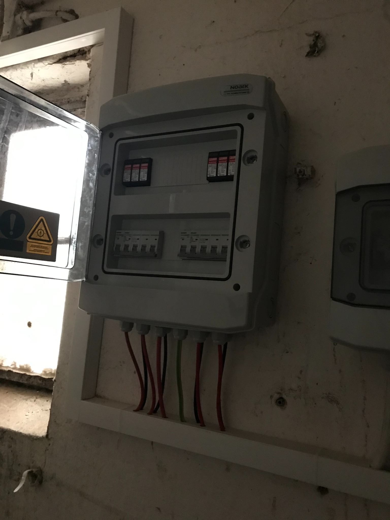 Szara skrzynka elektryczna z otwartymi drzwiczkami, zamontowana na ścianie. Widoczne bezpieczniki i przewody. Obok druga, mniejsza skrzynka. Ściana w tle jest lekko zabrudzona.