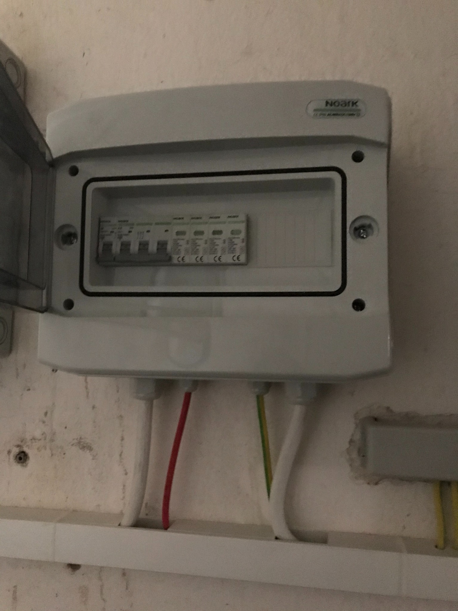 Otwarta skrzynka elektryczna Noark z widocznymi bezpiecznikami i przewodami w kolorach: białym, czerwonym i zielono-żółtym, zamontowana na ścianie.